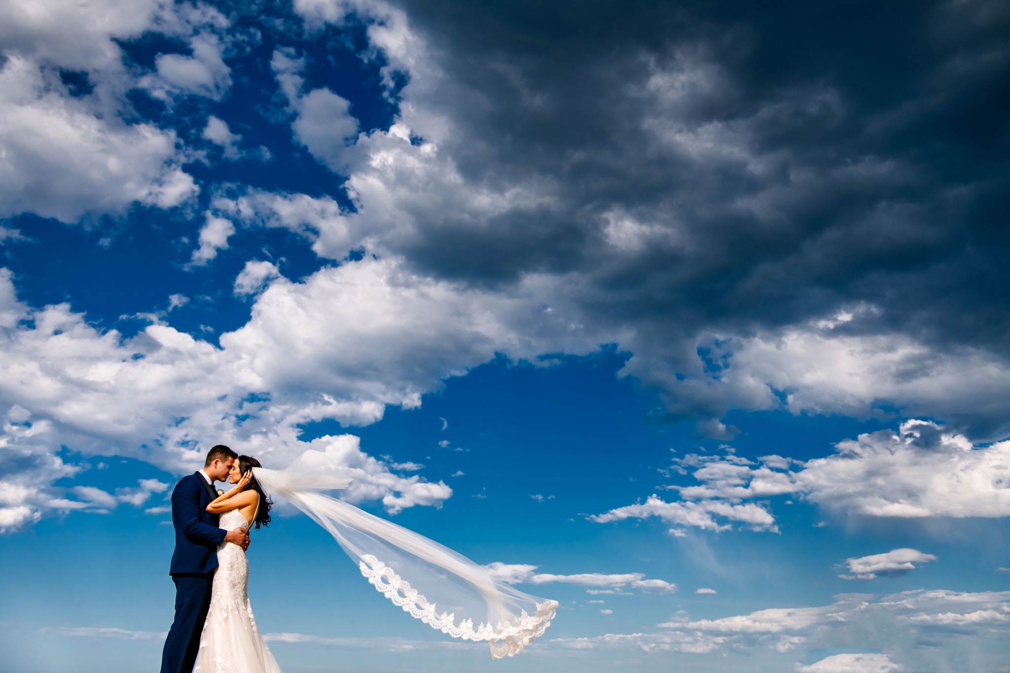 Newlyweds embrace with view of sky behind them as the bride's veil floats in the breeze