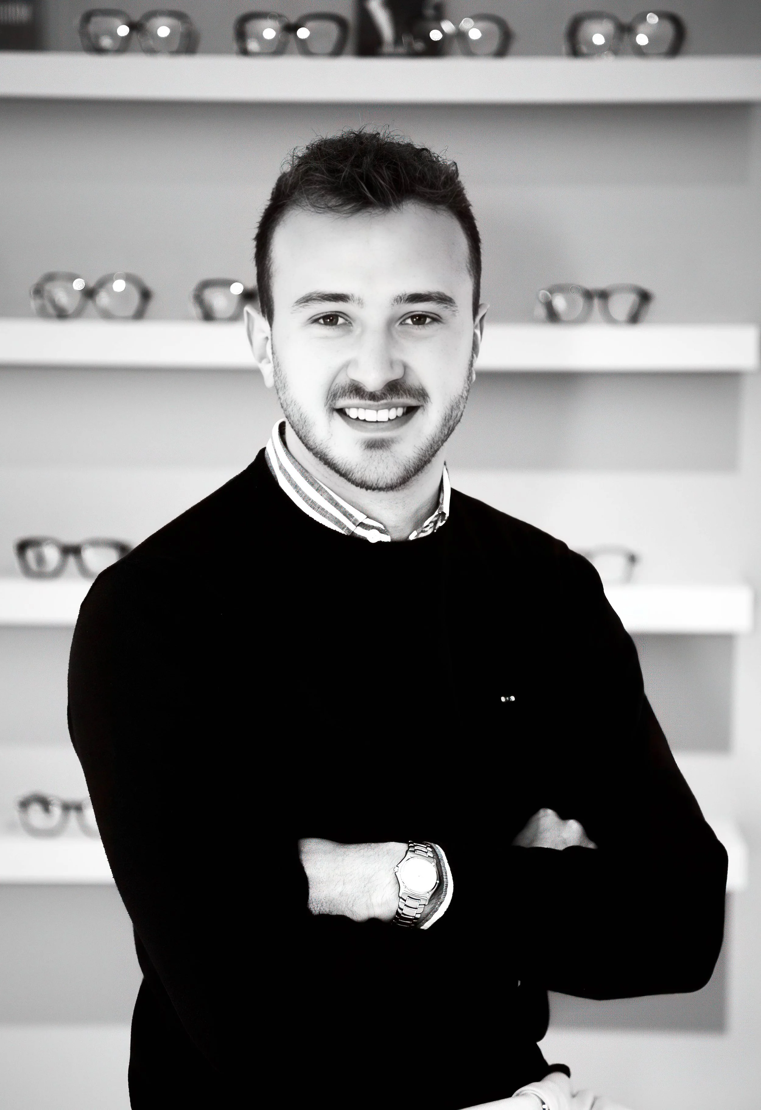Un jeune homme souriant avec une montre, portant un pull noir et une chemise, avec des lunettes en arrière-plan sur des étagères.