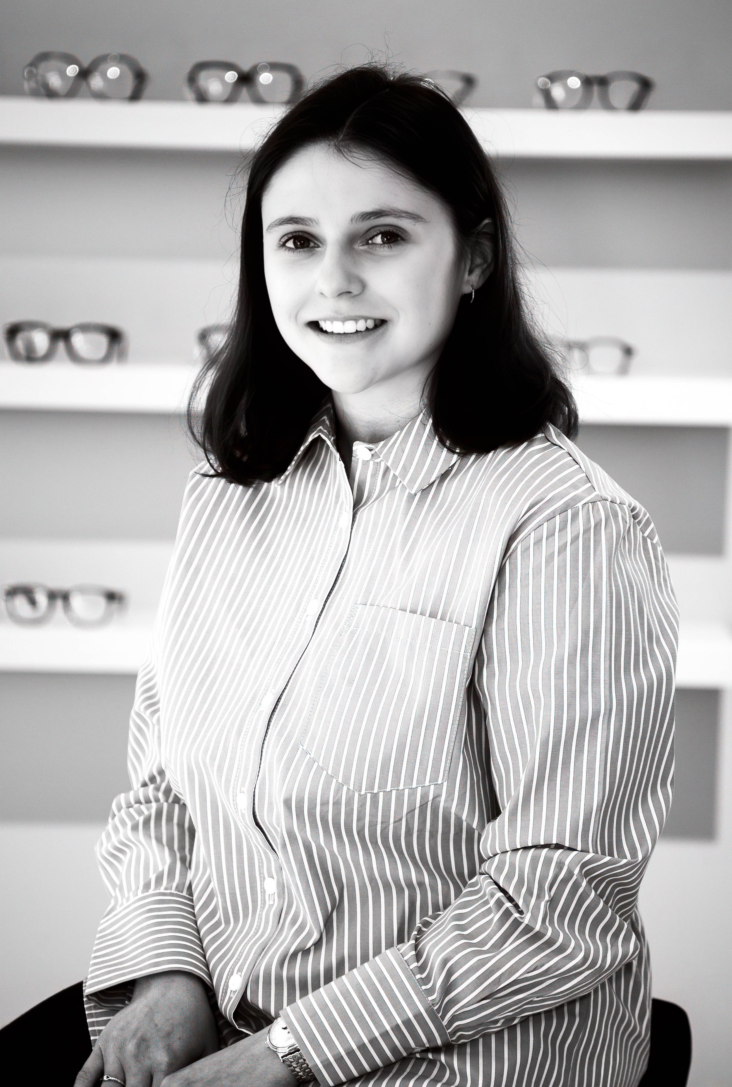 Jeune femme cheveux noirs, souriante, portant une chemise à rayures, assise dans un magasin de lunettes avec des montures de lunettes en arrière-plan.