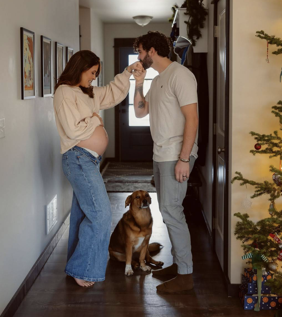 Pregnant woman and man sharing a kiss in a hallway, with a dog sitting between them and a decorated Christmas tree nearby.