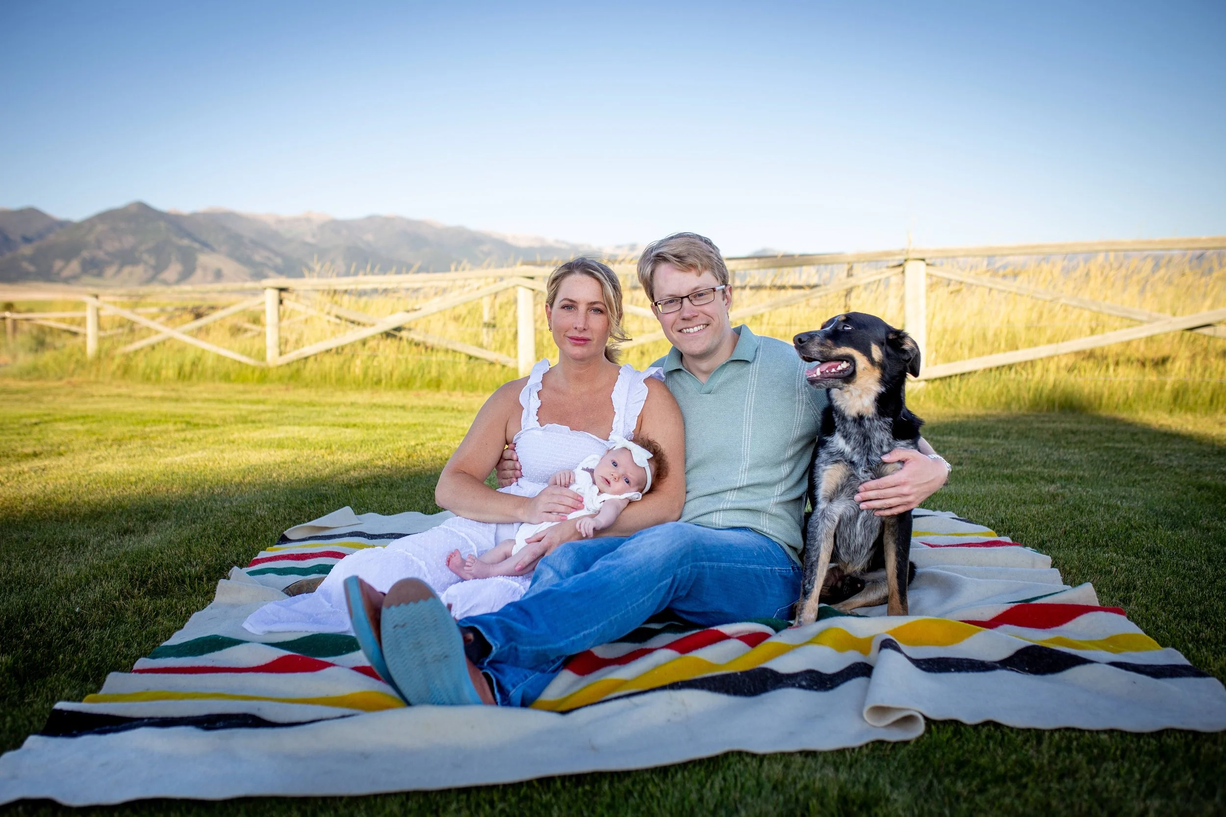 A family of four sitting on a colorful striped blanket outdoors. The mother is holding a baby, the father is sitting next to her with a dog in his lap, and mountains are visible in the background.