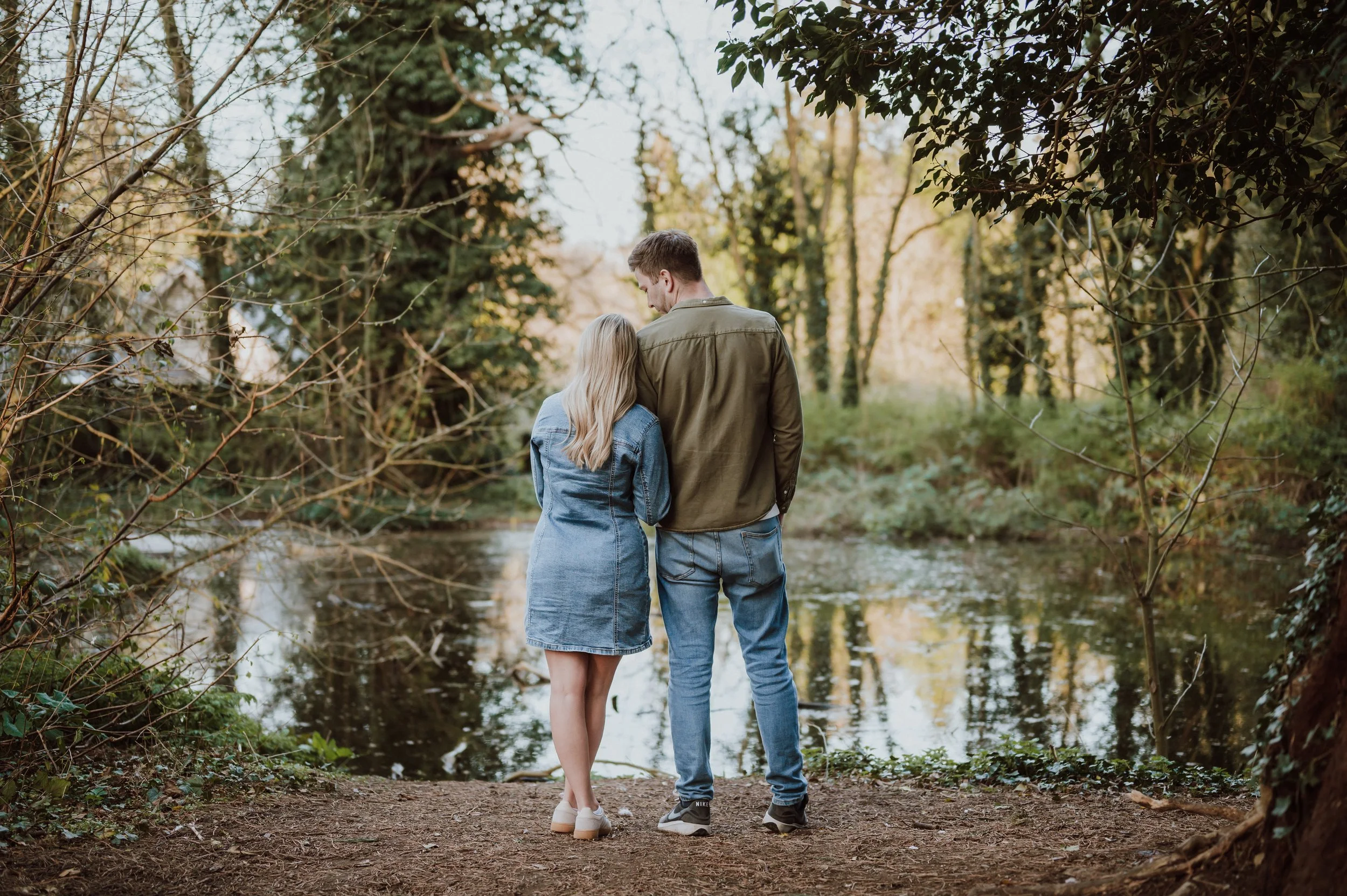 A young couple stands close together on a dirt trail near a creek, surrounded by trees with some autumn foliage, as they look at each other in a peaceful outdoor setting.