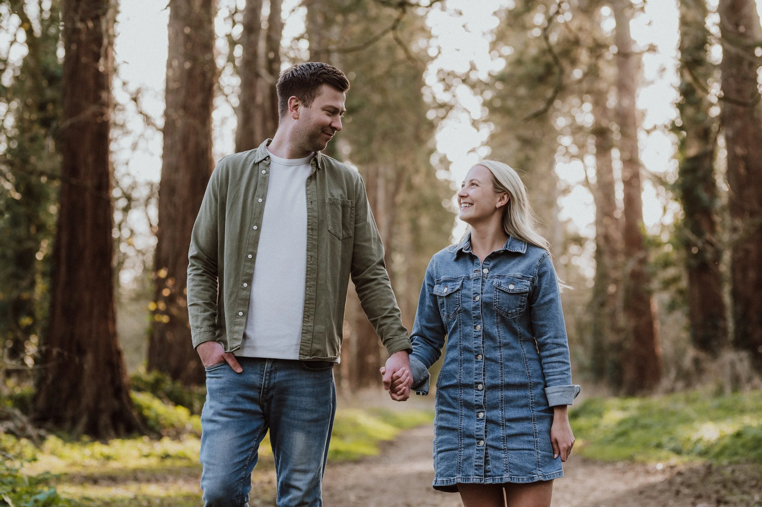 A couple holding hands walks through a forested area, smiling and looking at each other, surrounded by tall trees with sunlight filtering through.