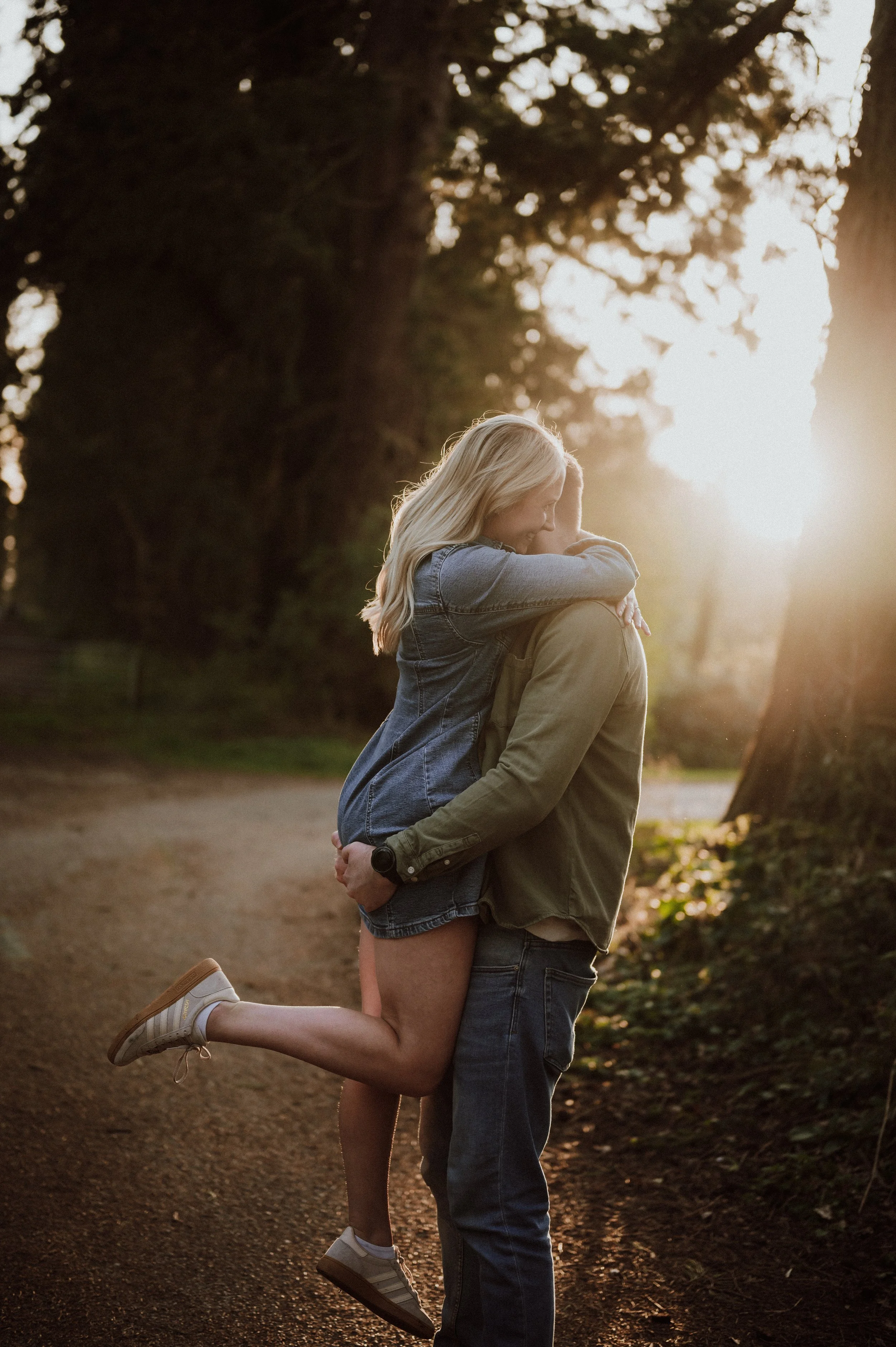 A couple embracing outdoors on a dirt path during sunset, with trees in the background.