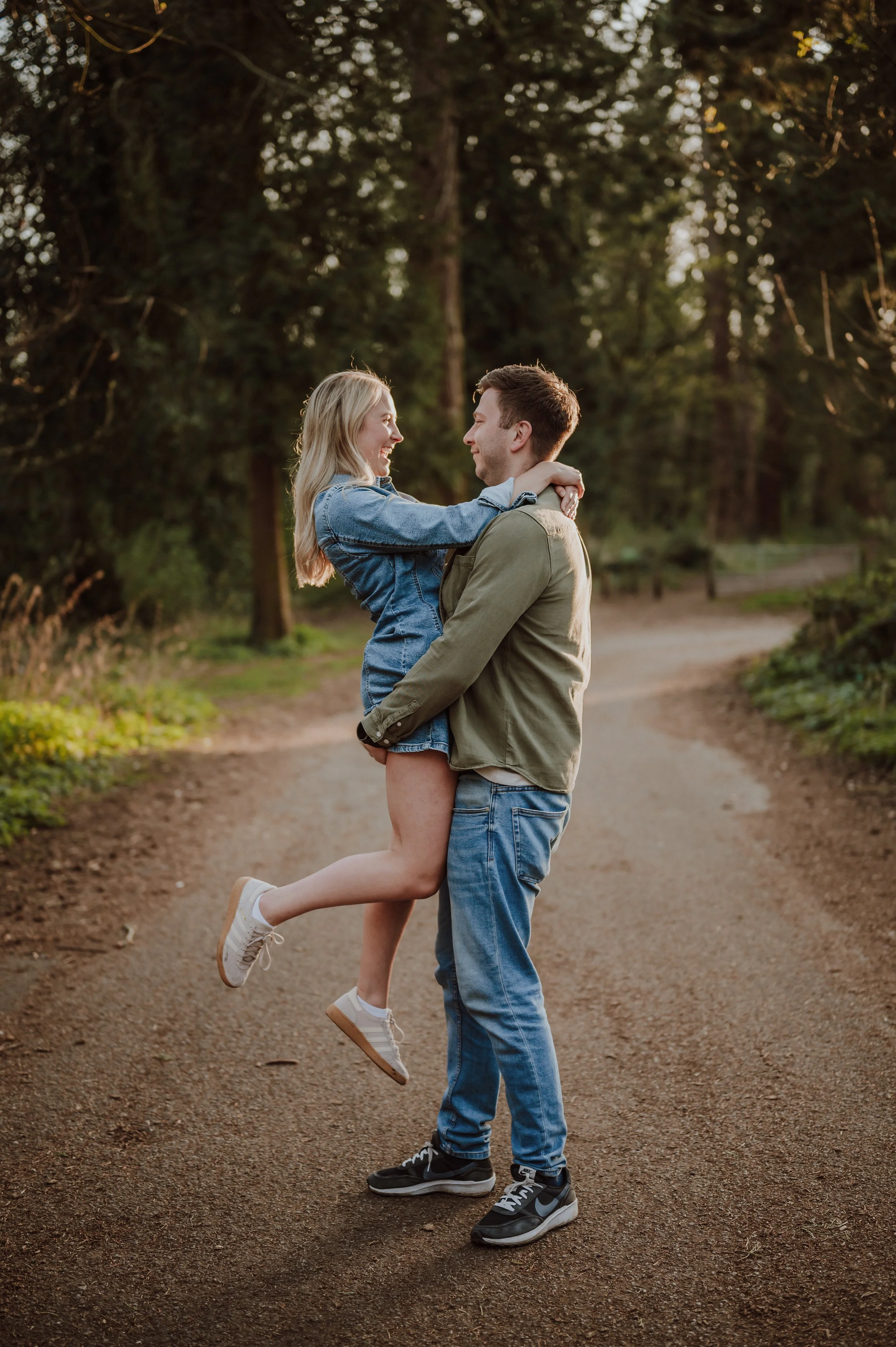 A young couple in casual clothing is outdoors on a dirt path in a wooded area, with the man lifting the woman while they look at each other and smile.