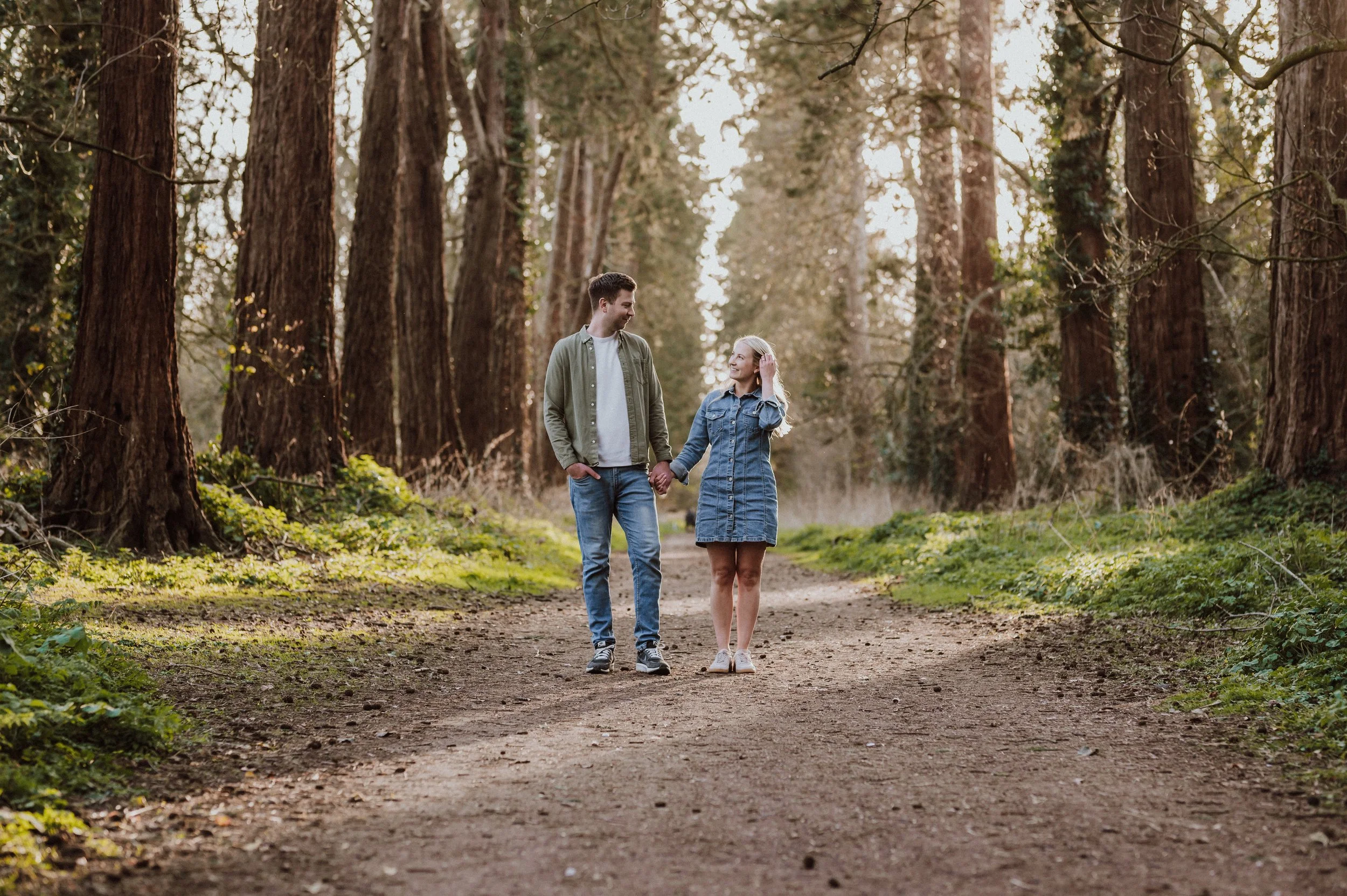 A young couple walking hand-in-hand on a dirt forest trail with tall trees on either side, during late afternoon or early evening.