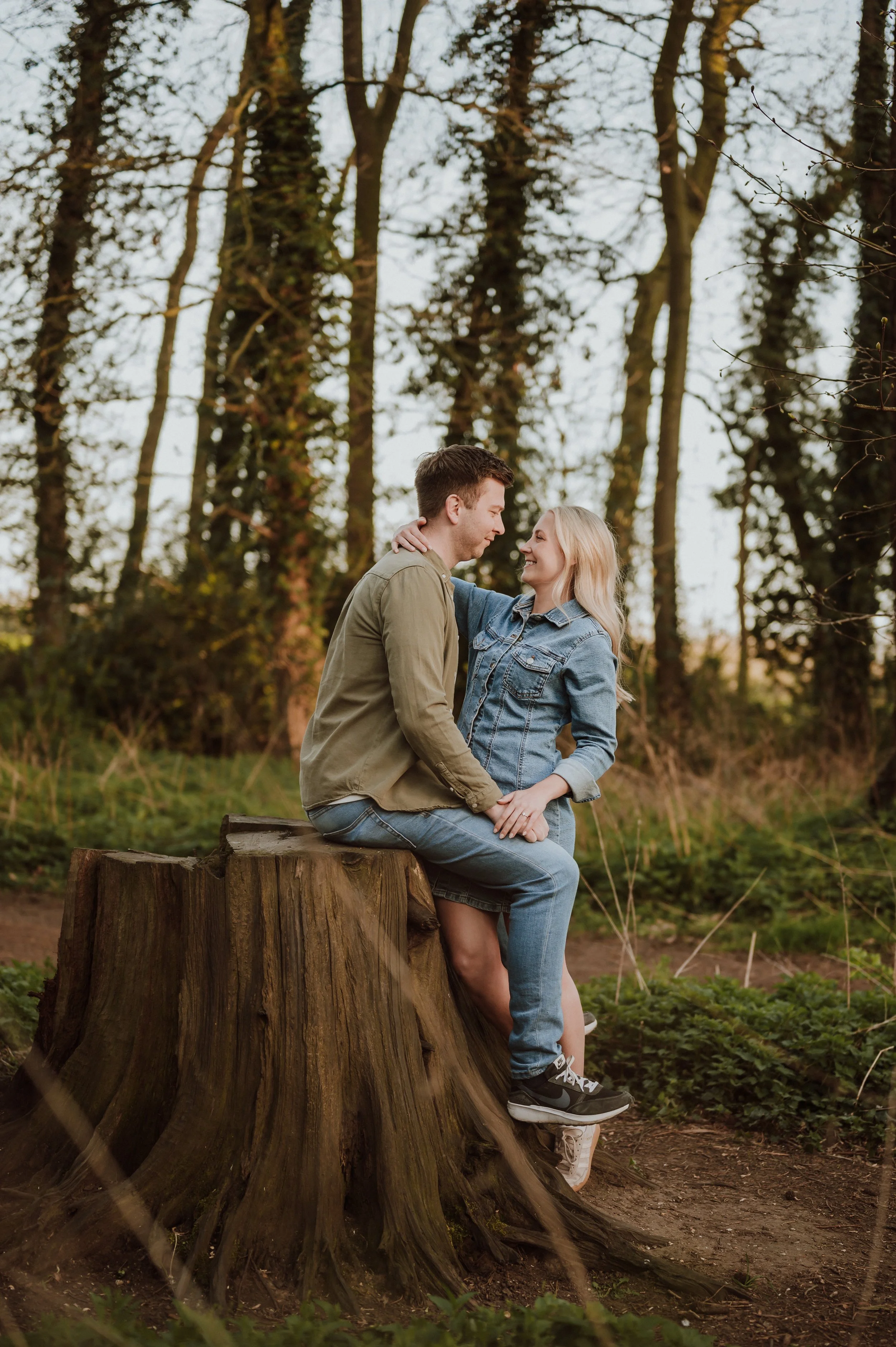 A young couple sitting on a tree stump in a forest, facing each other and smiling, with trees in the background.