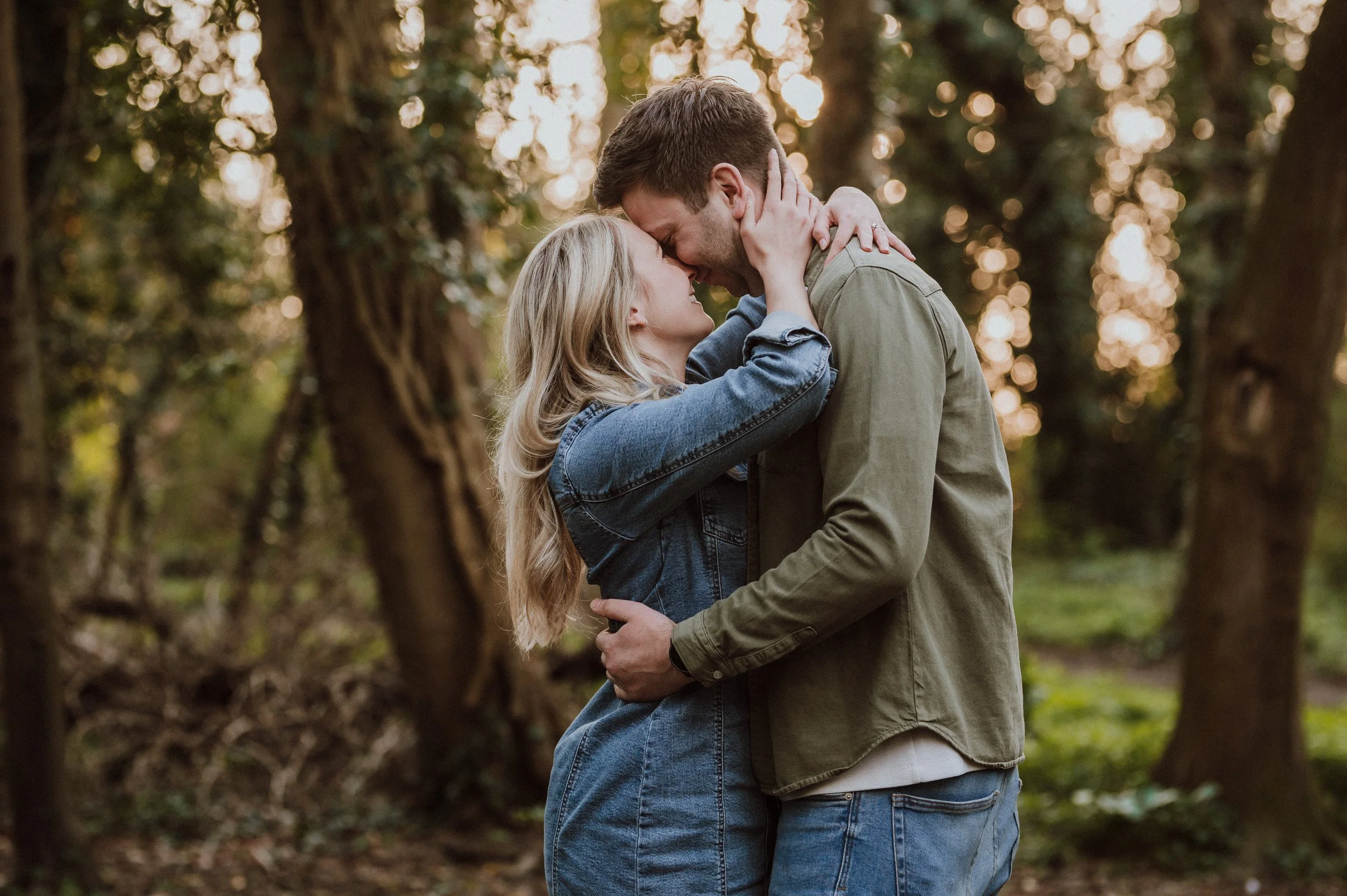 A couple embracing in a wooded forest at sunset, with the woman cradling the man's face and the man holding the woman around her waist.