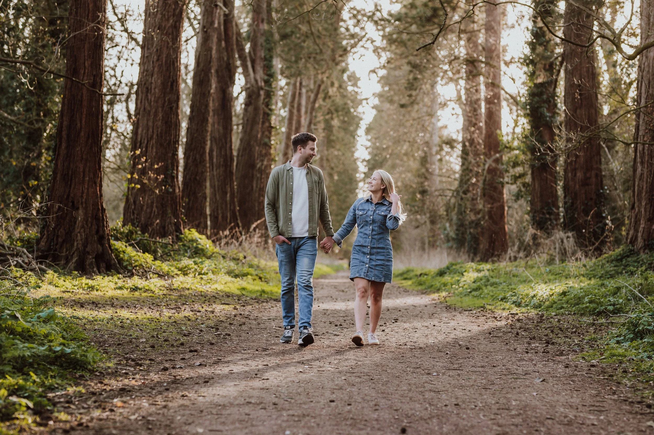 A young couple holding hands and walking through a forest trail during daytime, surrounded by tall trees and green foliage.