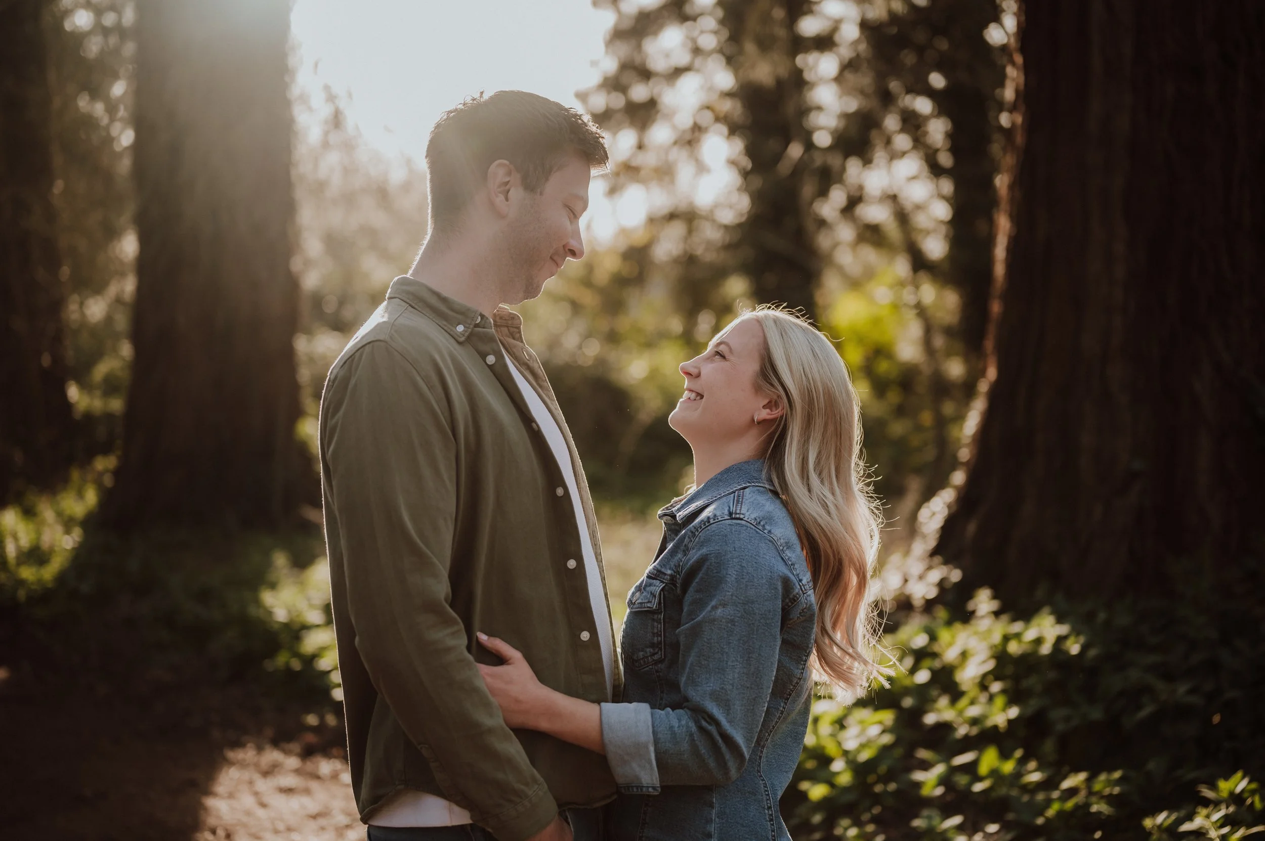 A couple standing close together in a forest, smiling and gazing into each other's eyes with sunlight filtering through trees.