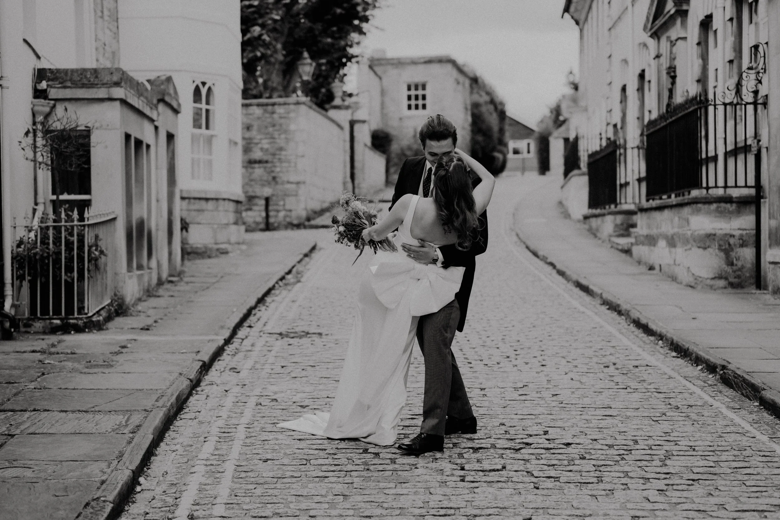 A black and white photo of a couple in wedding attire dancing on a cobblestone street, with buildings on either side.
