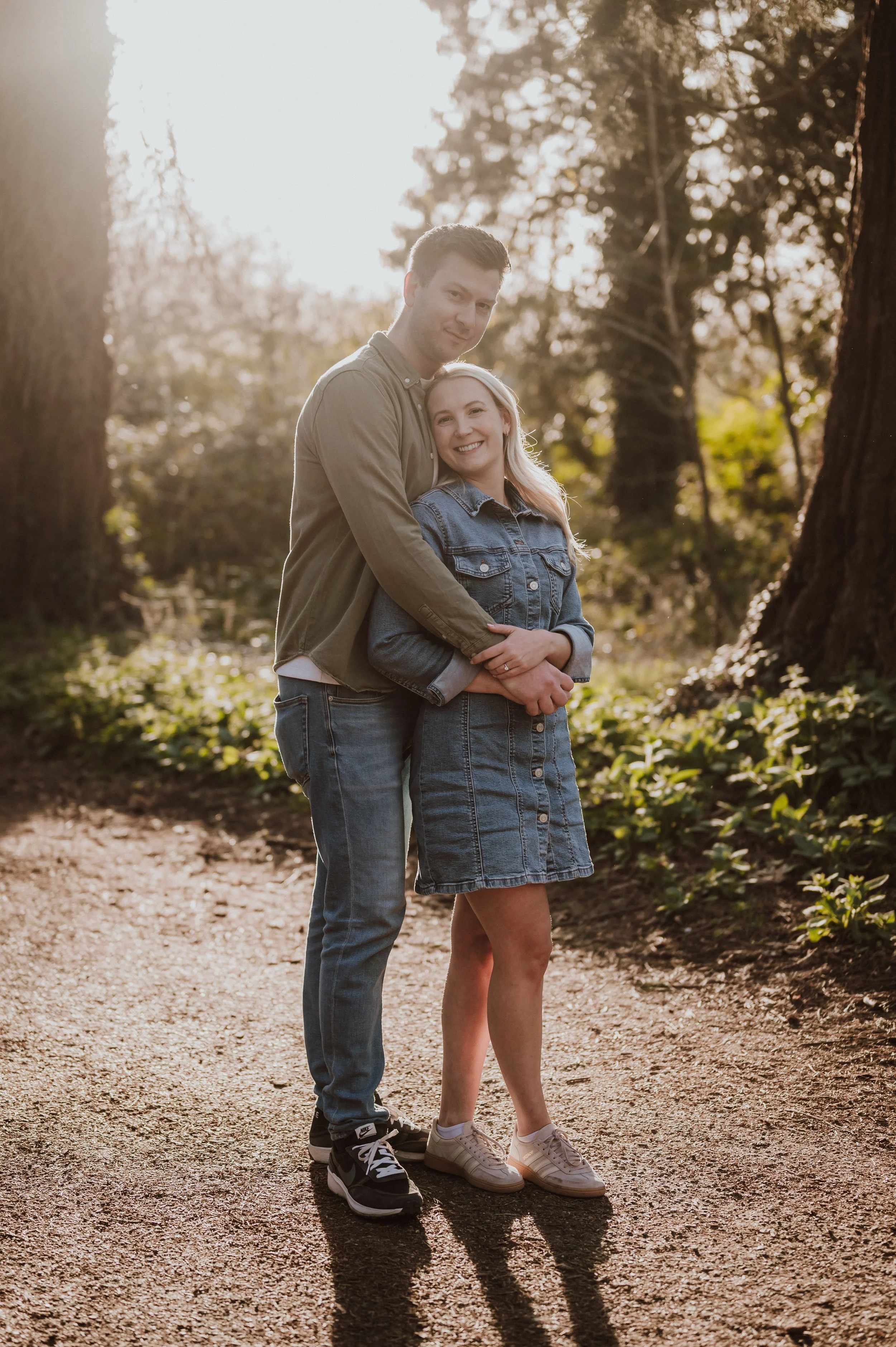 A young couple standing close together on a dirt path in a wooded area during sunset, smiling and embracing each other.