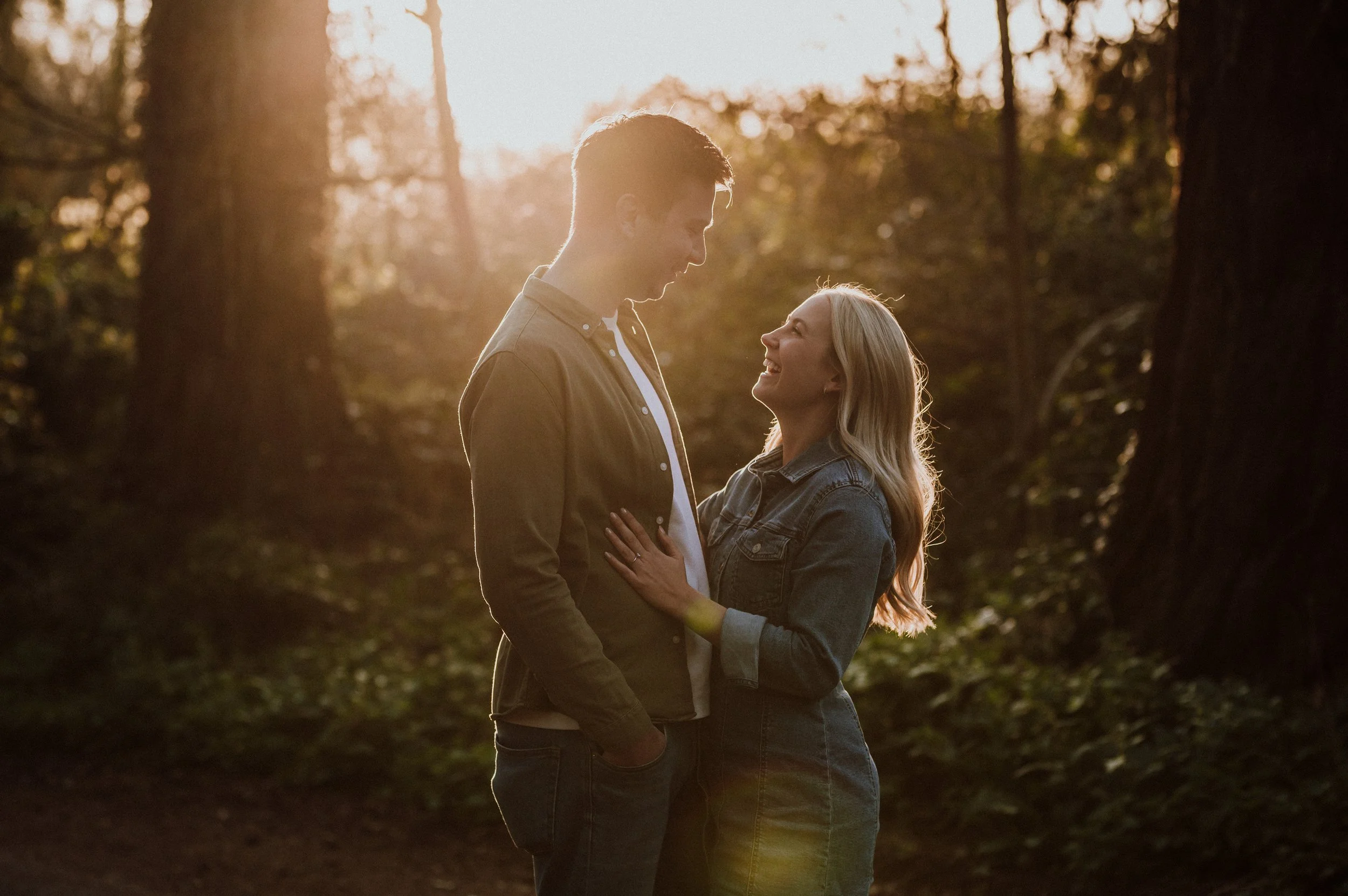 A couple standing close together in a forest during sunset, smiling at each other.