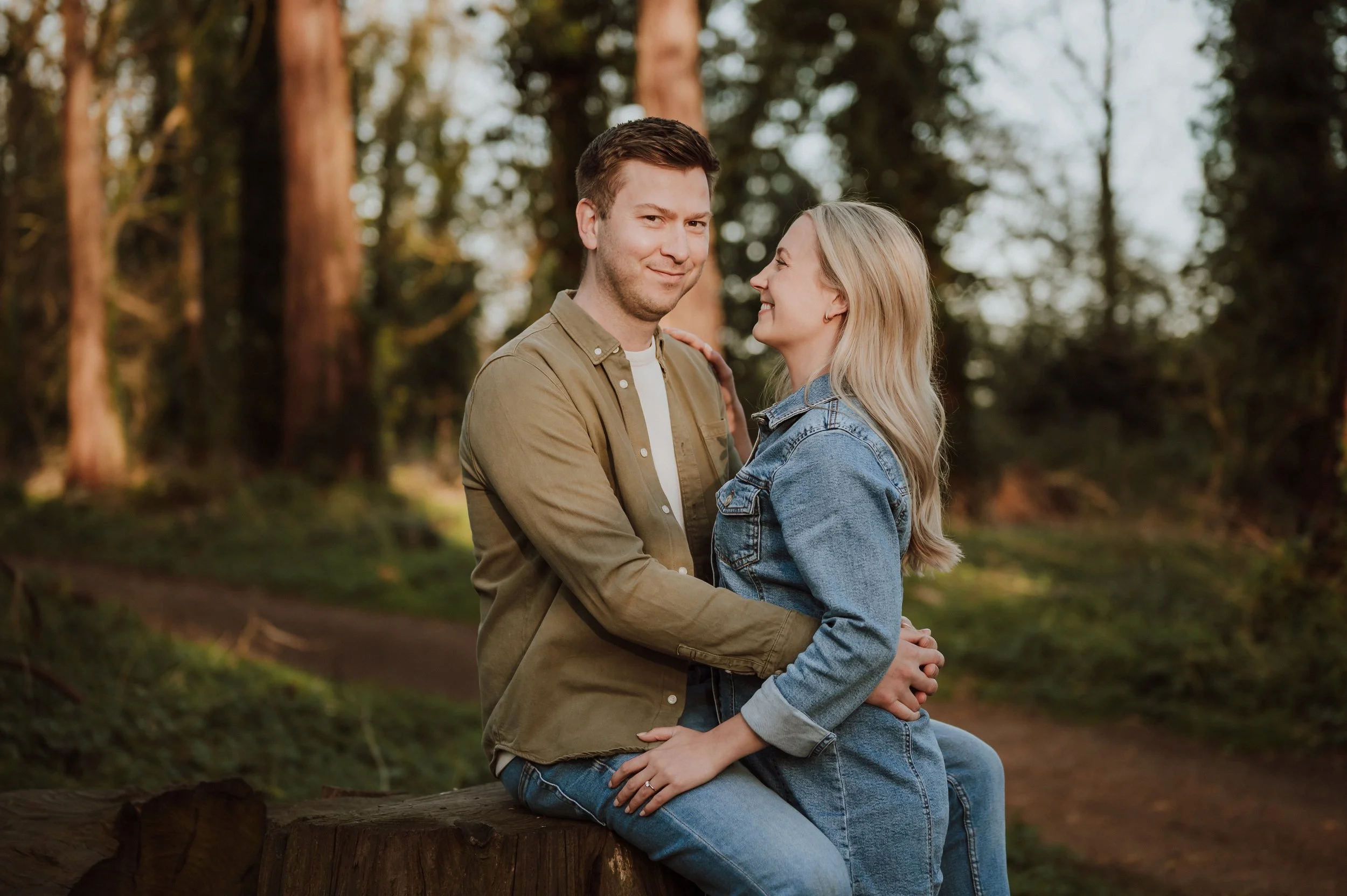 A couple sitting on a tree stump in a forest, smiling and embracing, with tall trees and sunlight in the background.