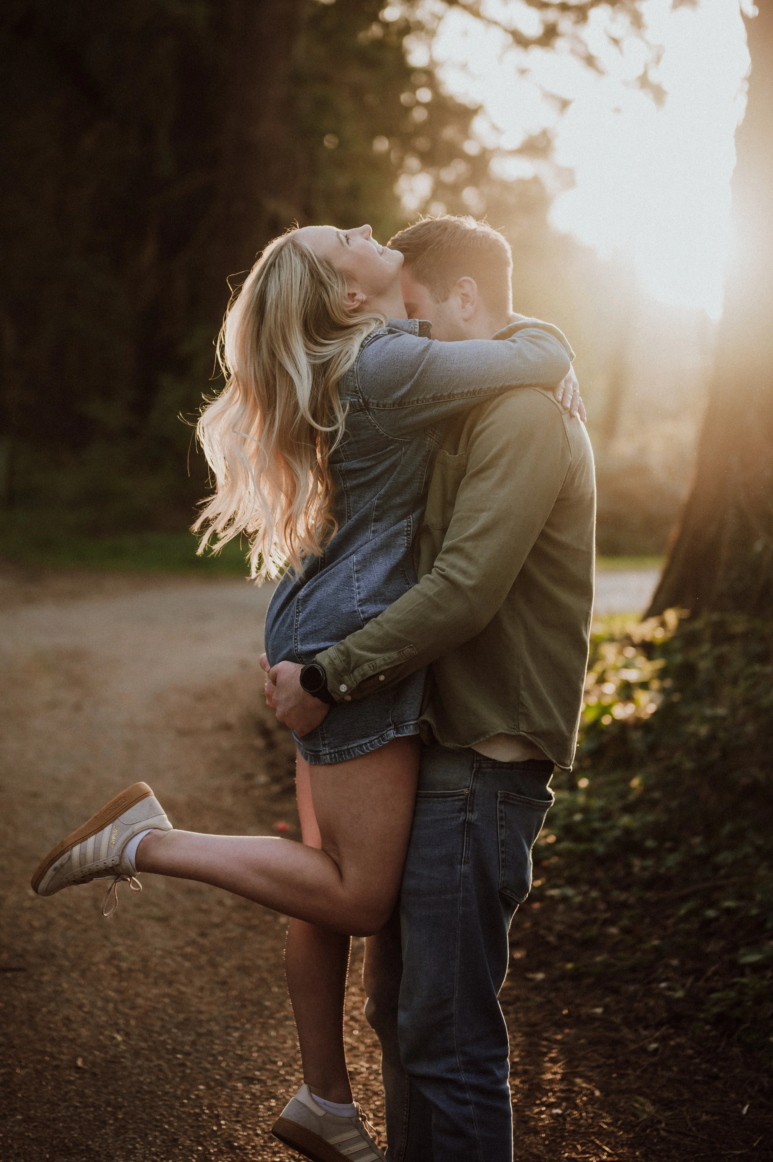 A couple embracing outdoors during sunset, with the woman lifting the man off the ground, both smiling. The woman has long blonde hair, wearing a denim jacket and shorts, and the man has short hair, wearing a green shirt and jeans.