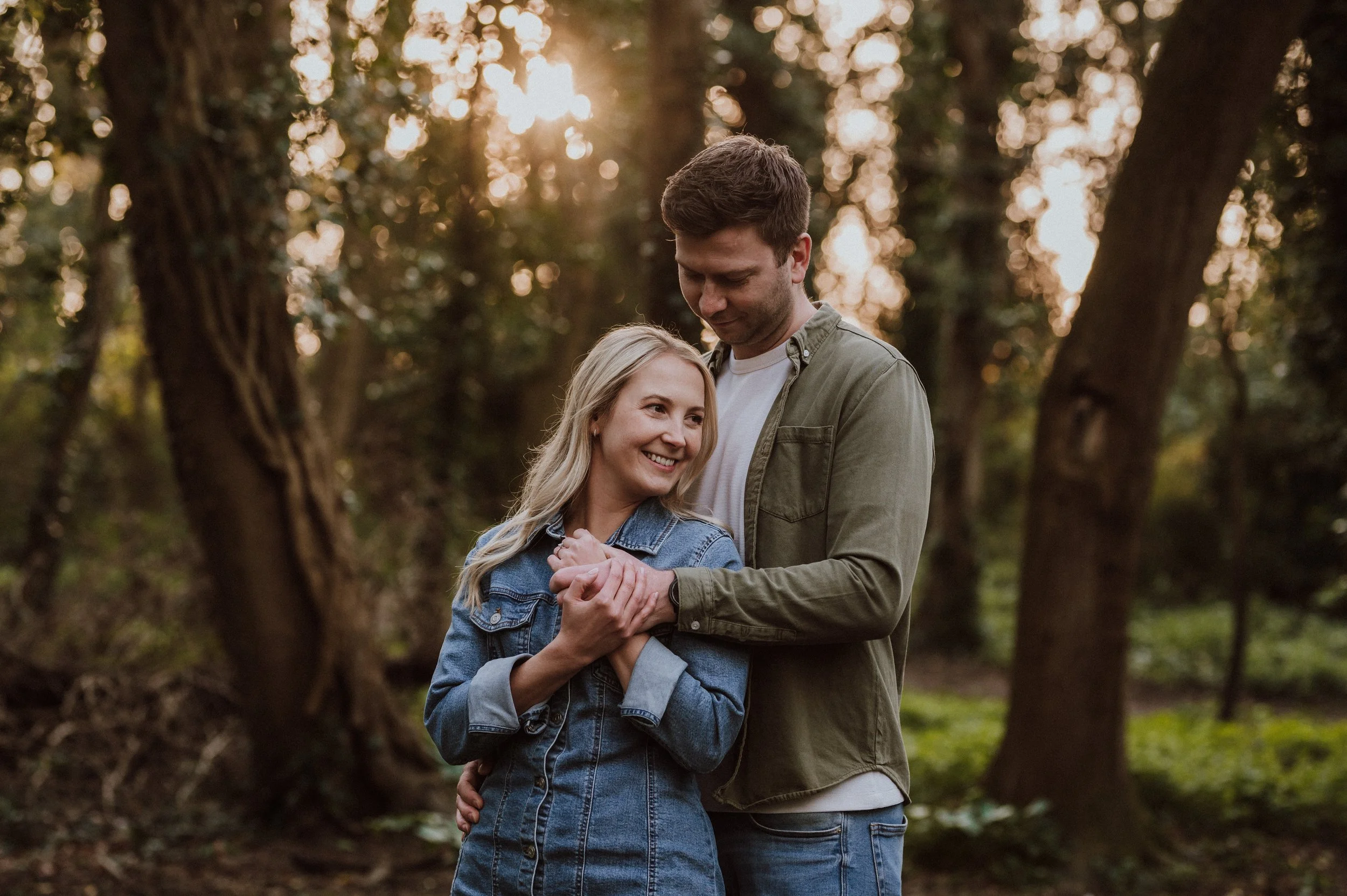 A couple standing in a forest at sunset, smiling and holding hands.