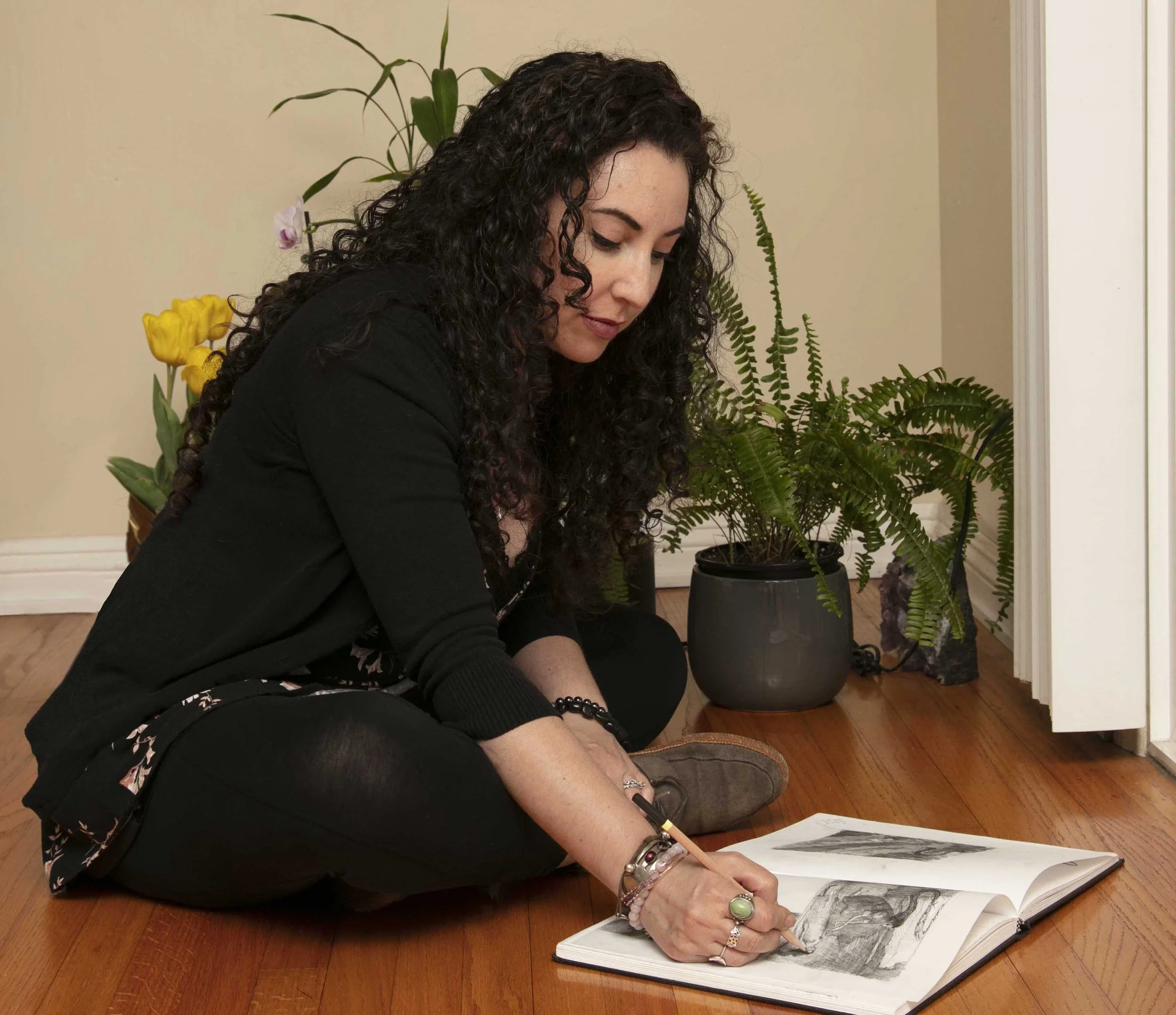 A woman with curly dark hair sitting on the hardwood floor, drawing in a sketchbook with trees in black and white. She is wearing a black jacket and surrounded by potted plants, including a fern and a yellow flower plant, in a room with beige walls.