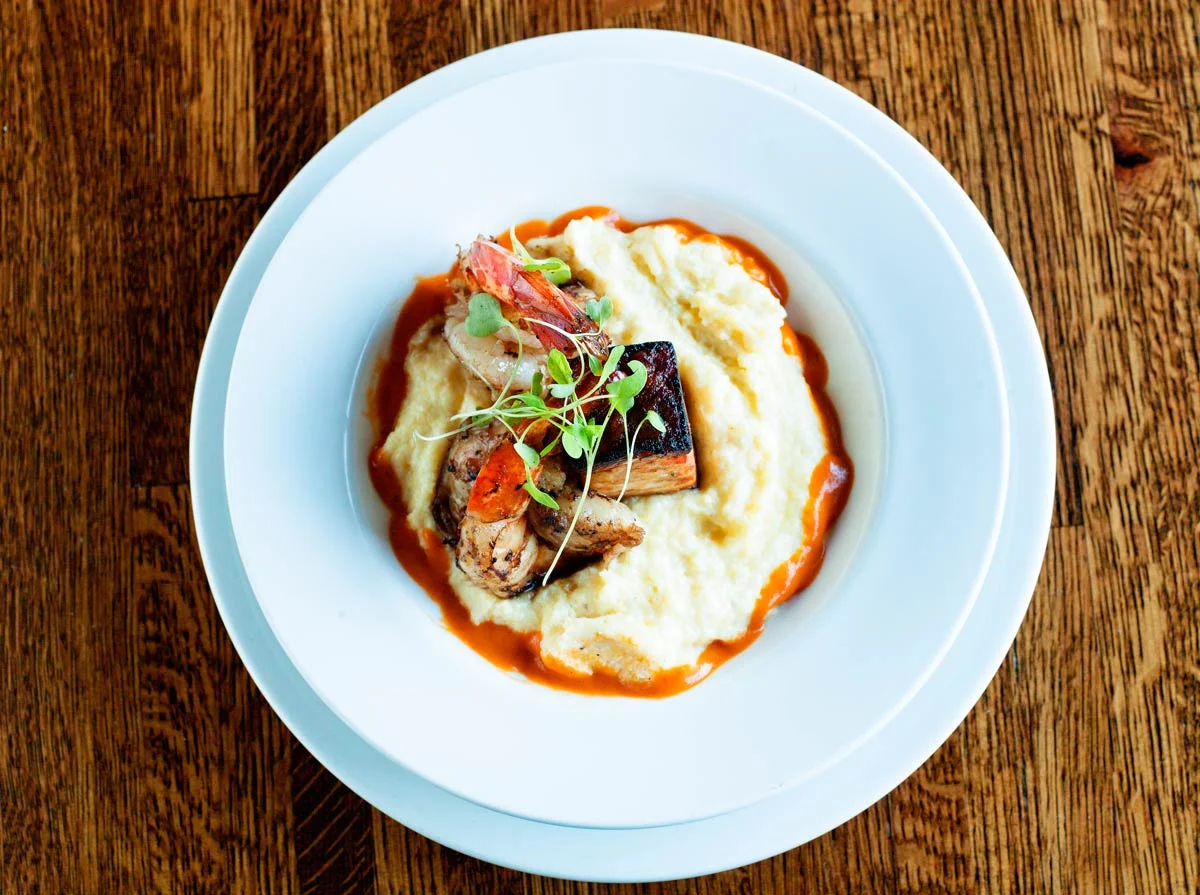 Plate of mashed potatoes with gravy topped with roasted meat, vegetables, and microgreens, on a wooden table.