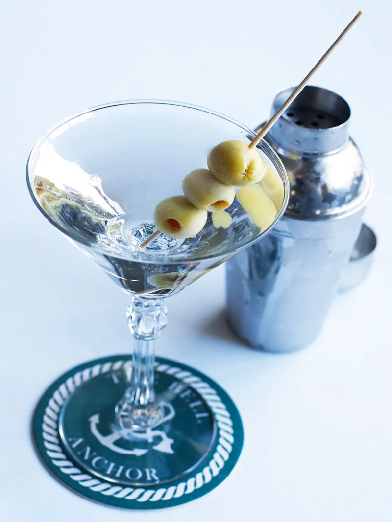 Cocktail glass with four green olives on a skewer, set on a coaster with an anchor symbol, near a cocktail shaker.