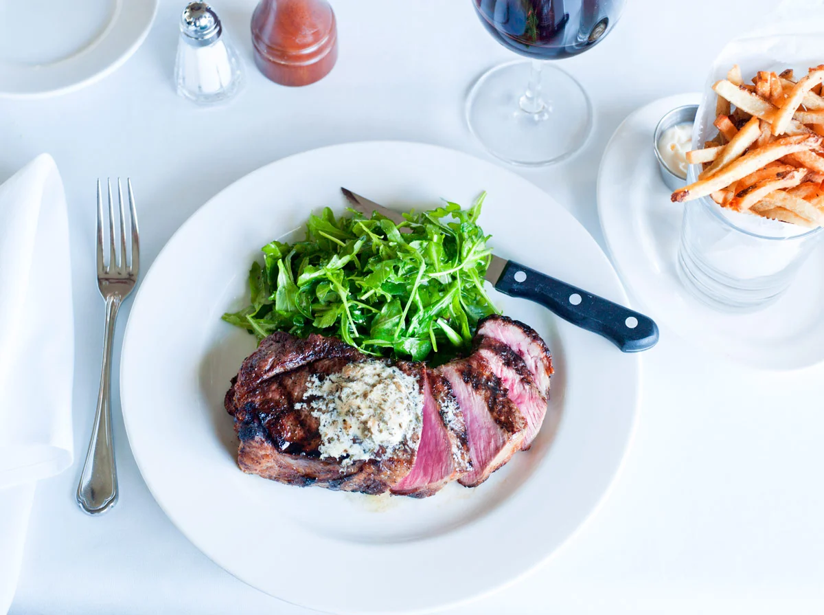 A white plate with a cooked steak topped with butter or cream, a side of arugula salad, and a knife. To the right is a glass of red wine and a glass of French fries in a glass container. There are salt, pepper, and a toothpick on the table, which is 