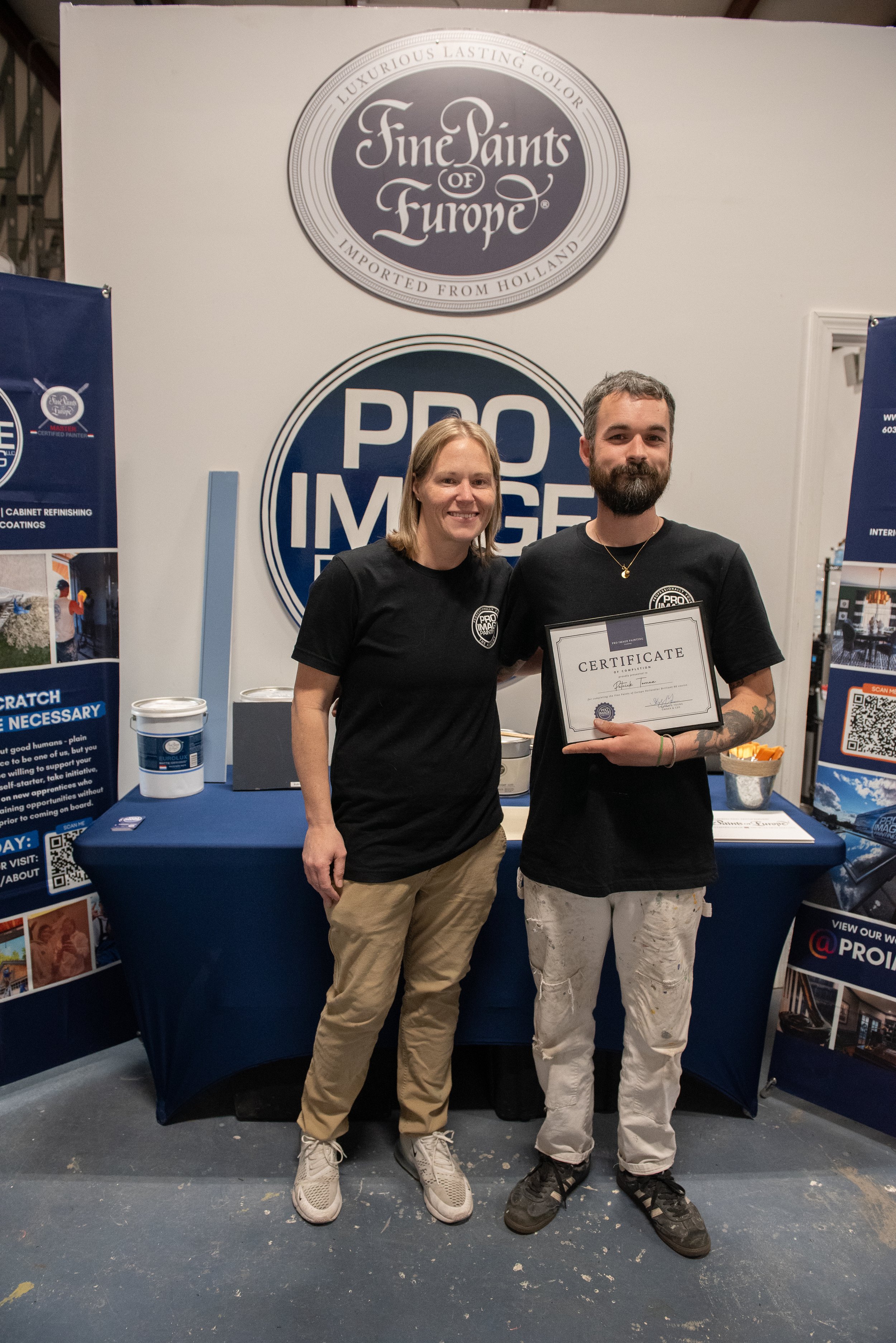 A man and a woman standing together at an event, both wearing black shirts. The woman is holding a certificate, and they are smiling at the camera. Behind them are banners with logos and text, and they are standing at a table with various items on it.