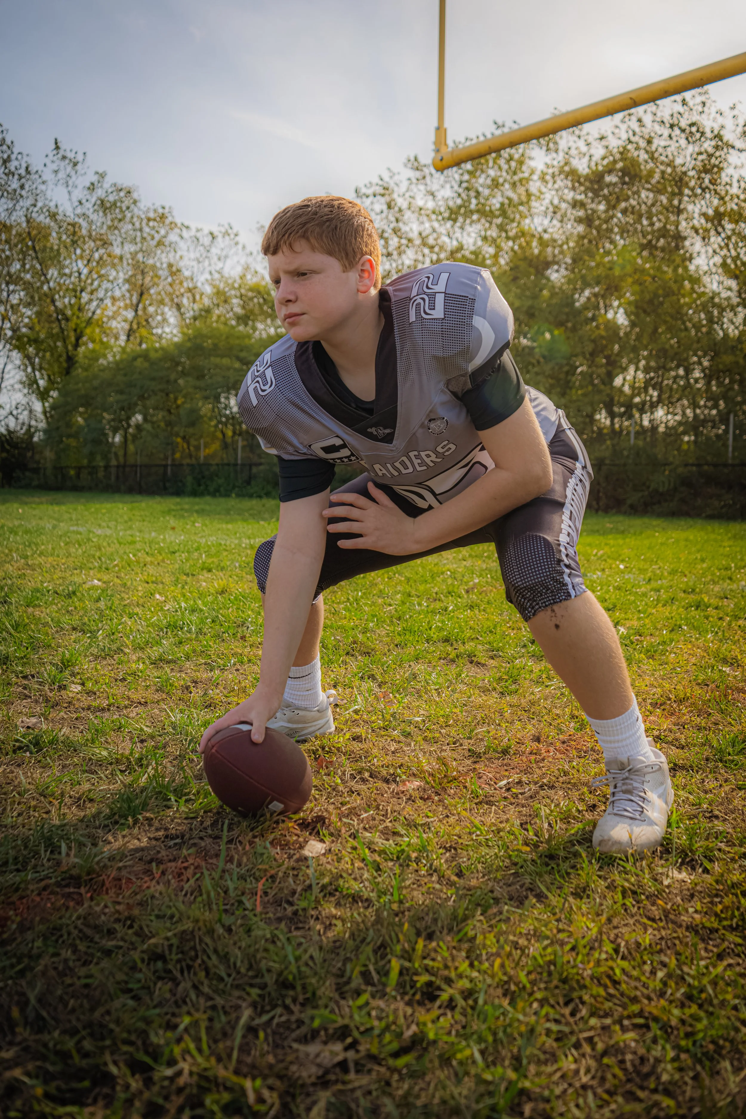 Young football player in grey and black uniform with number 22, crouched on the field, holding a football, preparing to make a play, during a game in the late afternoon.