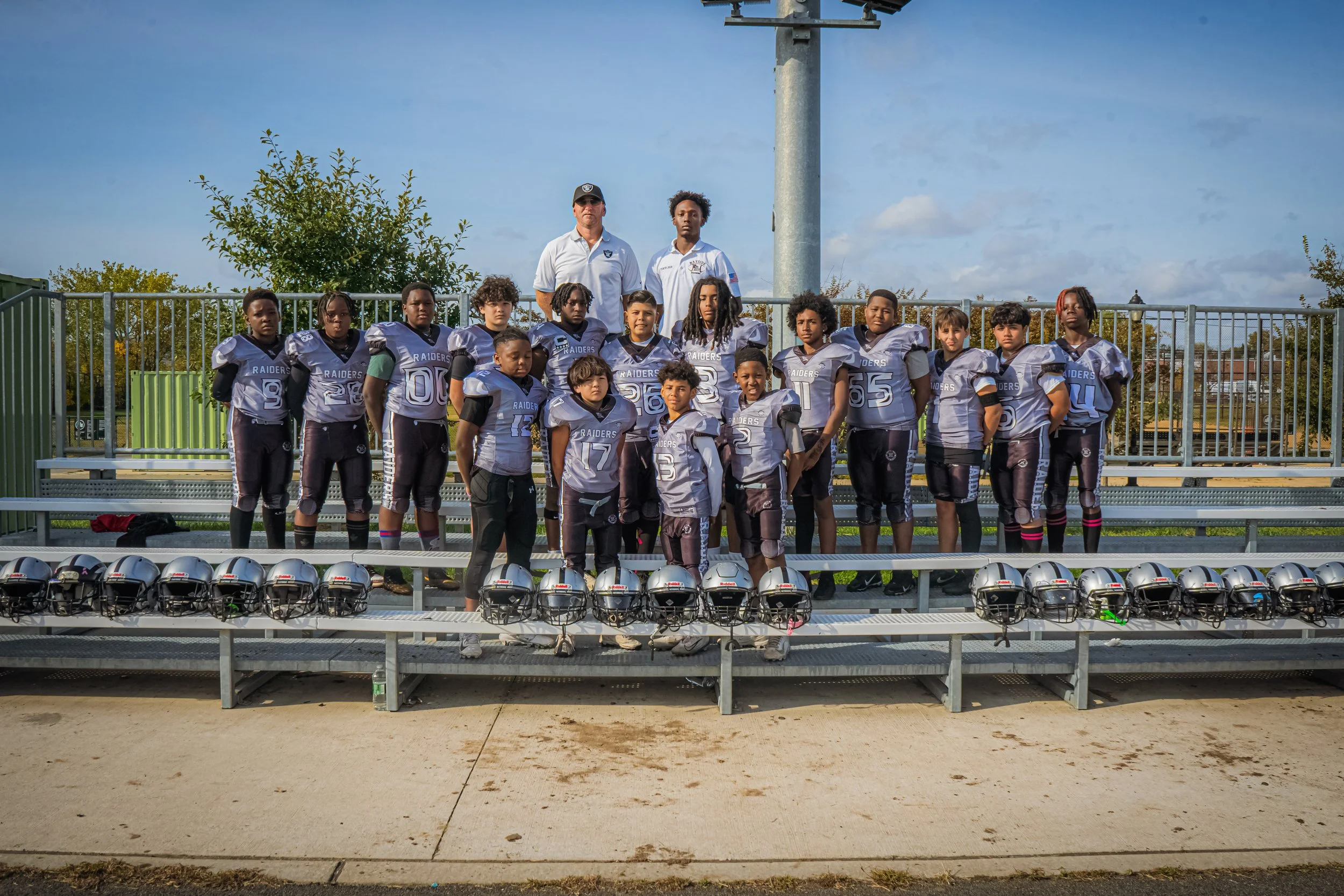 Youth football team with coaches standing on bleachers, wearing gray and black uniforms, helmets lined up on the ground in front, outdoor field with trees and blue sky.