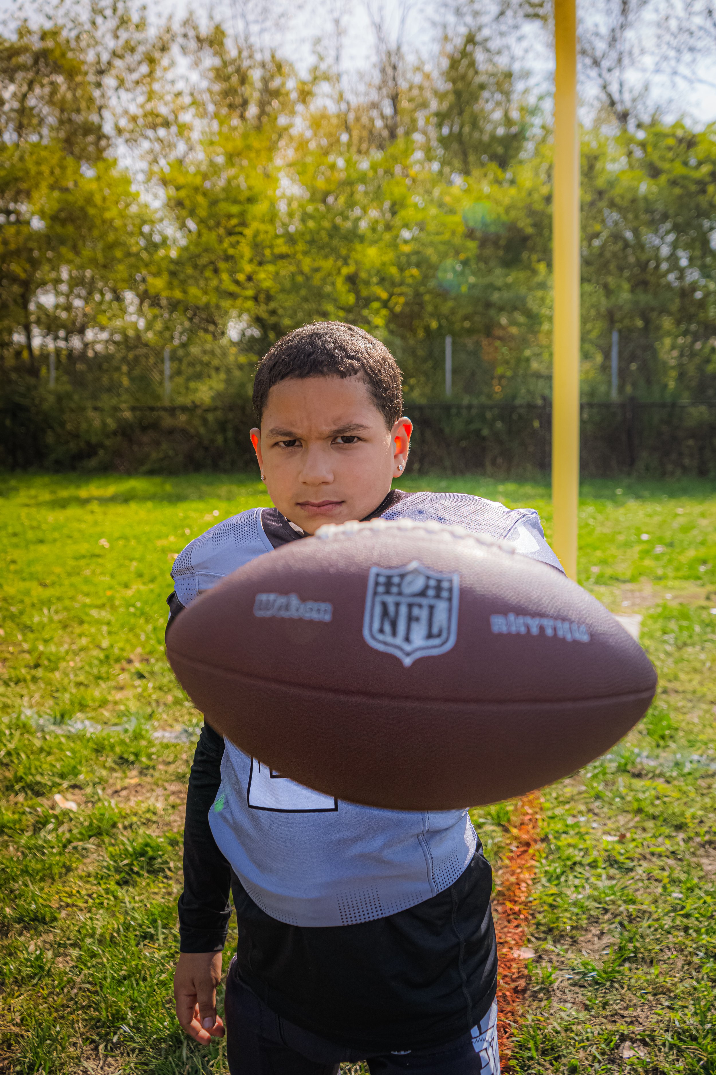 Young boy in a football jersey pointing a football toward the camera on a grassy football field with trees in the background.