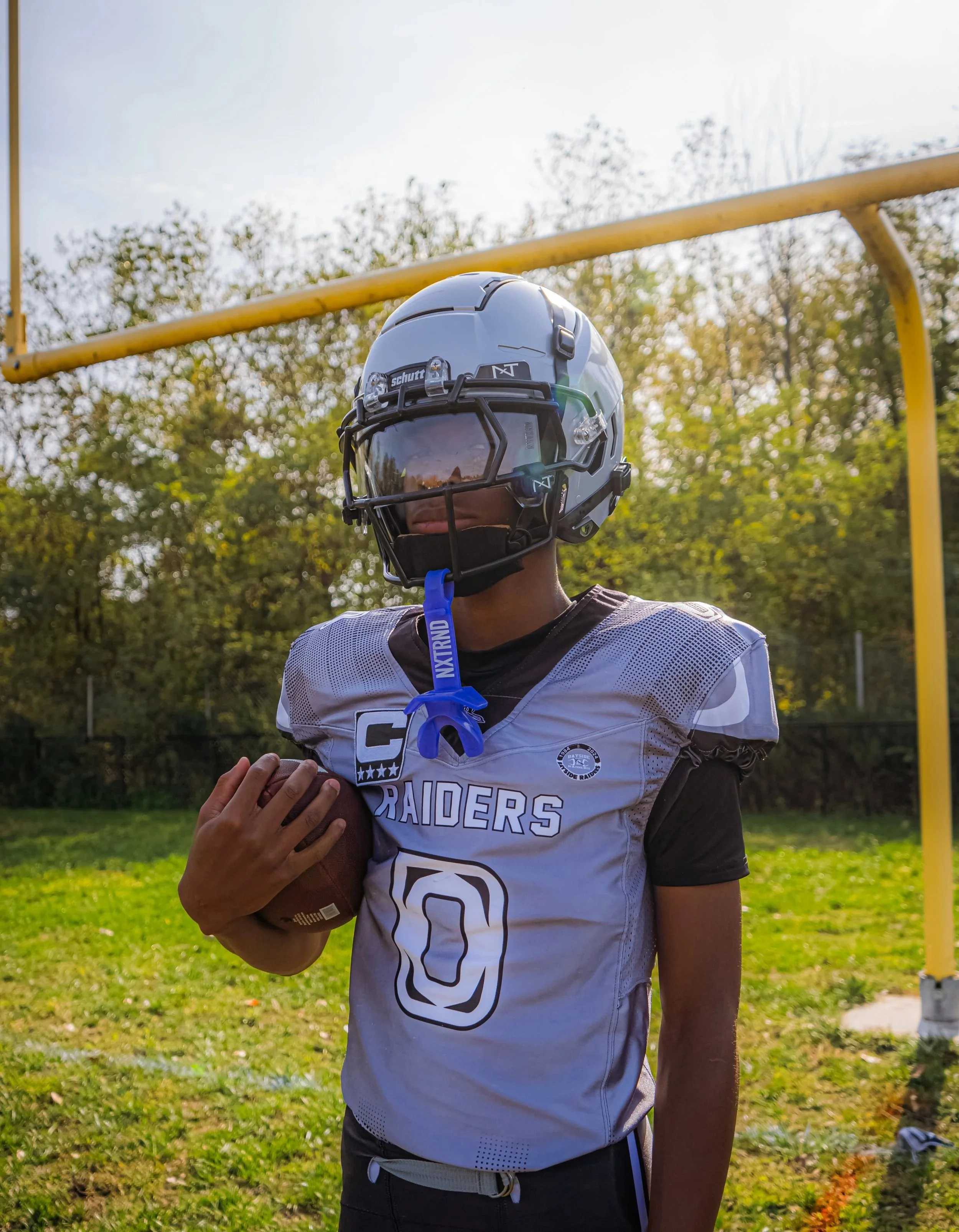 A young football player in a uniform holding a football on a grassy field, wearing a helmet with a face visor, with trees and a sunny sky in the background.