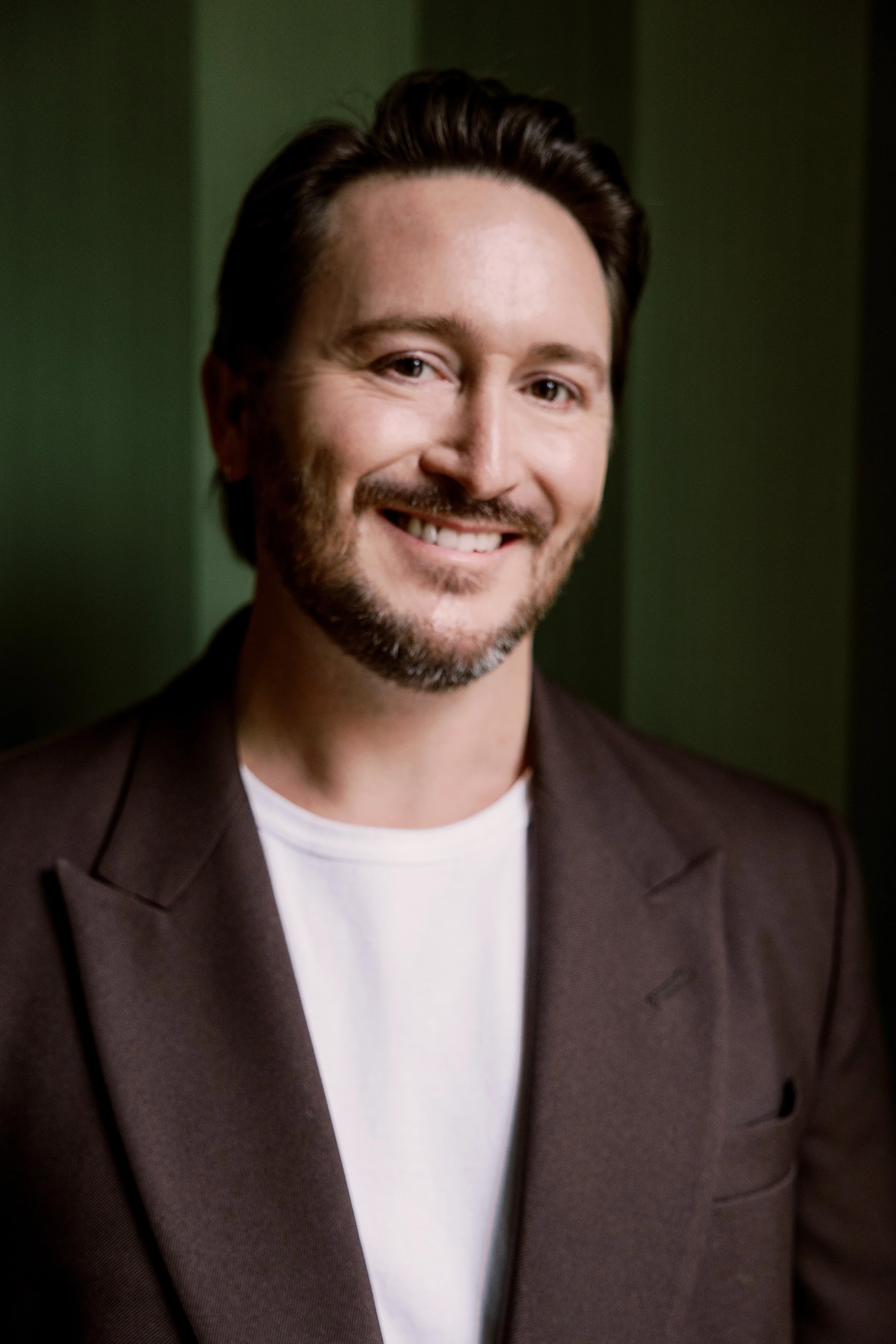 Close-up of a smiling man with dark brown hair, beard, and mustache, wearing a brown blazer and white T-shirt, against a dark green background.
