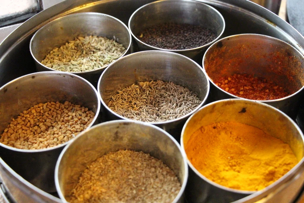 A spice box like this is a staple in any Indian home. These seven spices are some of the most common in Indian cooking. Not all boxes are the same, but they usually have some of the same set of ingredients. In this box the spices are, from the top going clockwise, mustard seed, red chili powder, turmeric, mystery spice, fenugreek seed, fennel seed, and center is cumin seed.