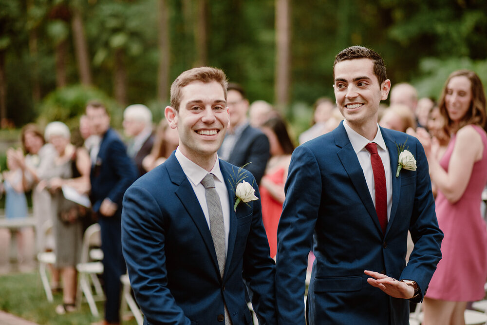  Two grooms walking down the aisle together. Vegan wedding at the Norfolk Botanical Gardens, Norfolk, VA. Rose garden and plant inspired wedding on the first day of Pride month. Sarah Mattozzi Photography. 