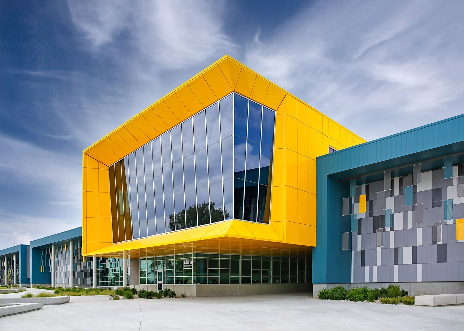 Low-angle architectural photography of a modern commercial building featuring a vibrant yellow geometric facade and grey textured paneling against a clear blue sky, captured by MANICPROJECT in Salt Lake City 