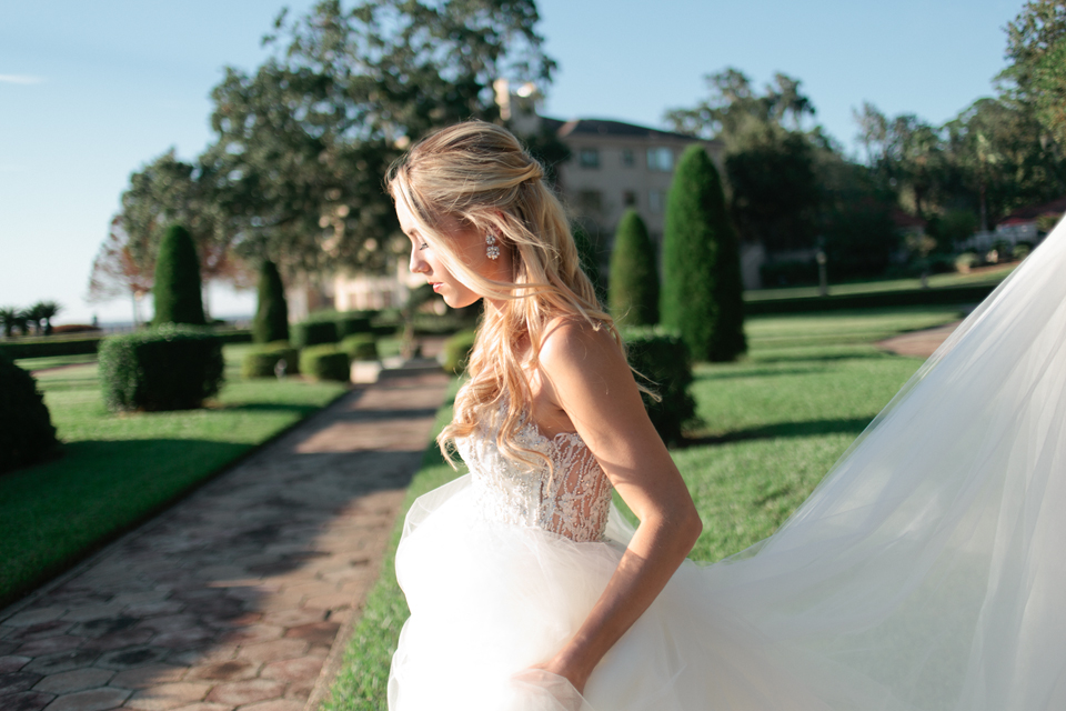 Picture of a bride walking and holding her Calvet Couture bridal gown.  She has long curls and is at the Epping Forest Yacht Club in Jacksonville, Florida