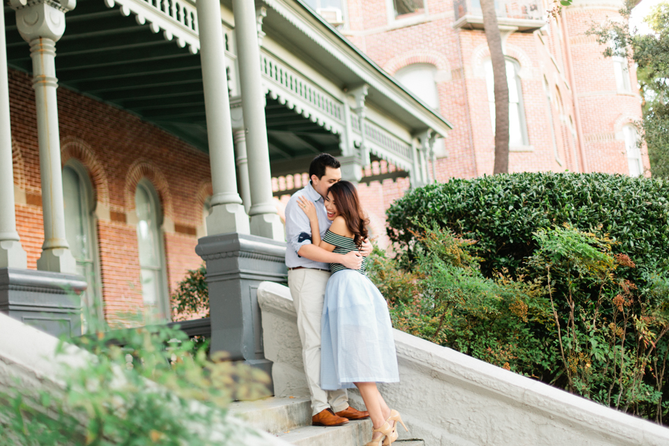 Picture of a couple on the steps of the Henry B. Plant Museum.  They are embracing, he is kissing her cheek, she is smiling.  This is an engaged couple.  