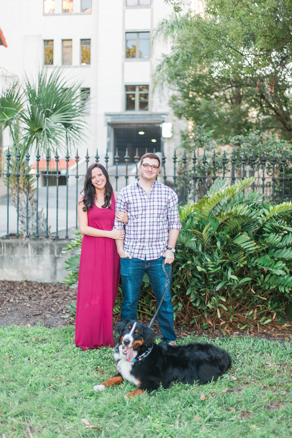 Image of an engaged couple standing together, linking arms, with their Bernese Mountain Dog downtown Orlando, Florida.  They are standing on grass in front of a black iron gate.
