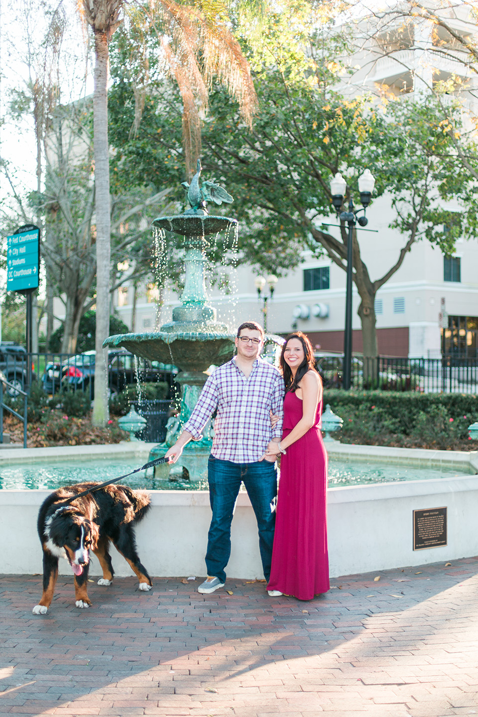 Picture of an engaged couple standing together in front of a fountain downtown Orlando with their Bernese Mountain Dog.