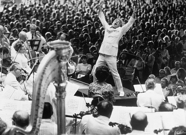 Leonard Bernstein and orchestra on a stage before thousands of people - he is wearing a white suit, arms outstretched and howling like a rock star
