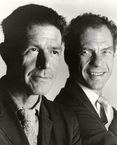 John Cage with Merce Cunningham slightly behind him, looking back and smiling at a serious Cage.