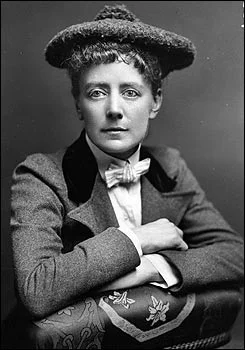 Ethel Smyth in a tweed coat and hat, arms folded over the back of a chair.