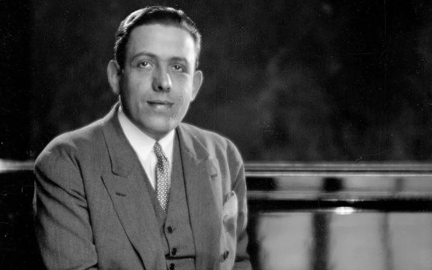 Poulenc in a suit under soft light with smoke rising from an unseen cigarette. 