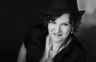 Catherine Laub in a fedora and tie with earrings, looking up at the camera and smiling.
