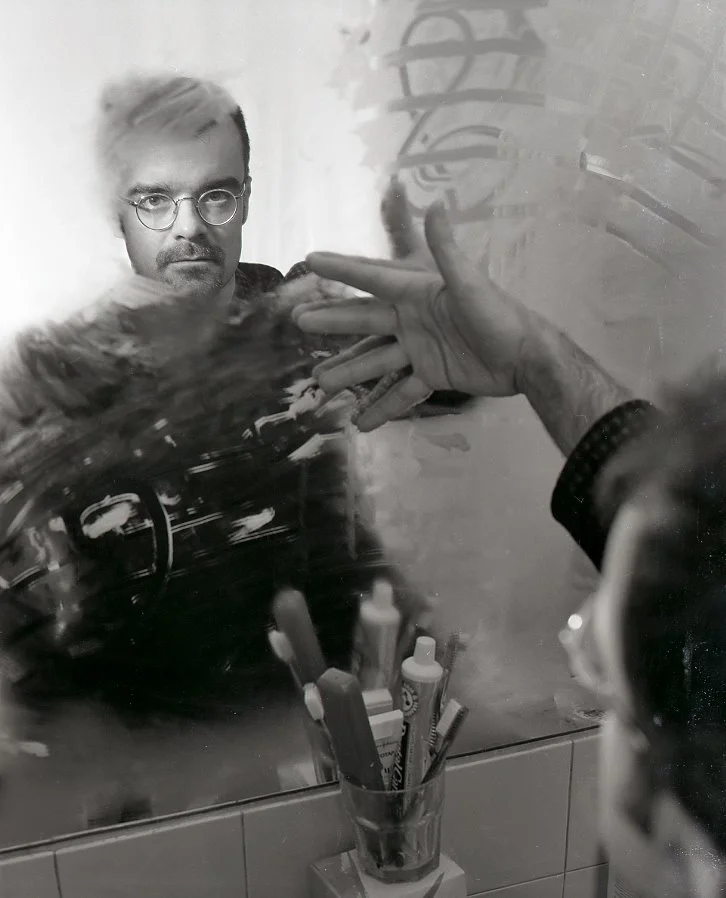 A yound Rodney Sharman looking at himself in a foggy bathroom mirror with a musical staff drawn in the condesation.
