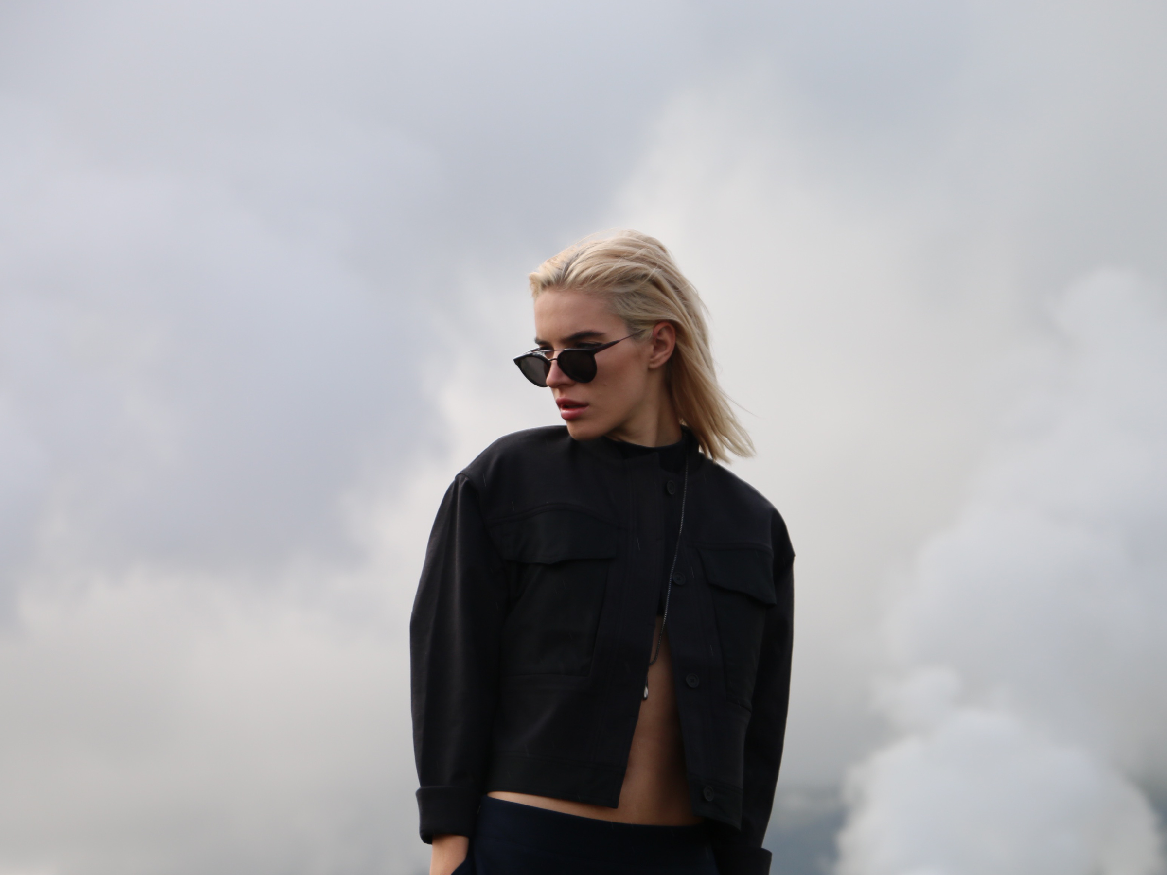 Chloe is wearing GIAGUARO FEMMENA Sunglasses by RETROSUPERFUTURE Cropped Flight Jacket + Front of a Line Skirt by Kit and Ace, JERN X ANTIQUED STEEL Pendant by Vitaly