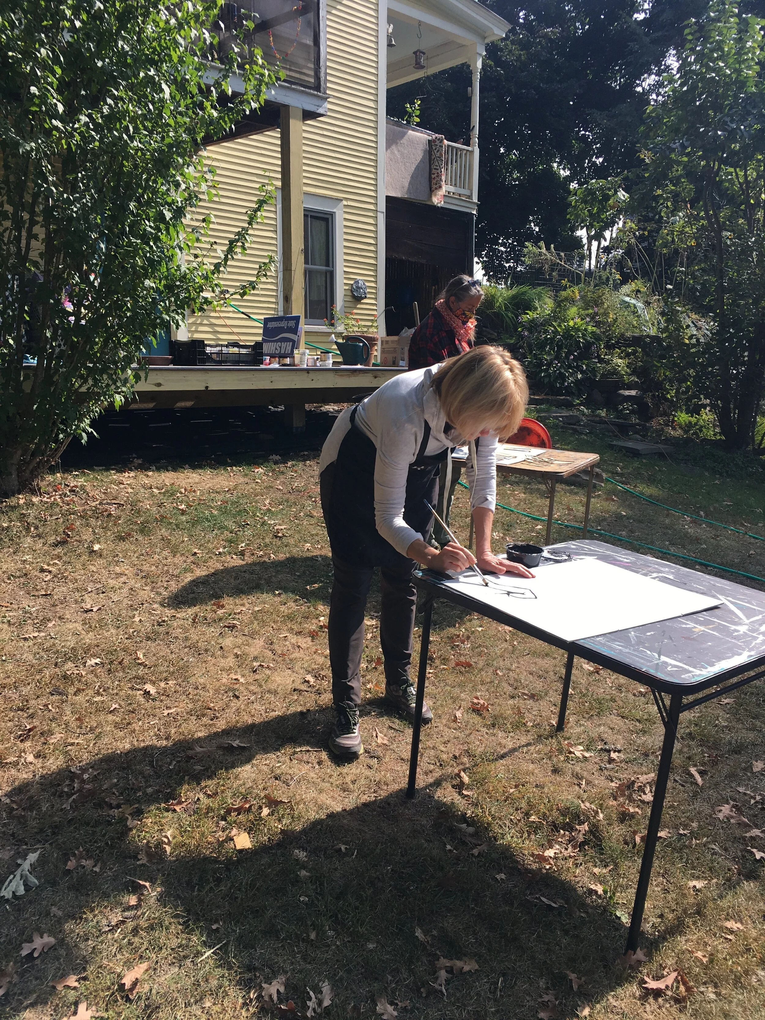 Two masked people making signs on card tables outside yellow house