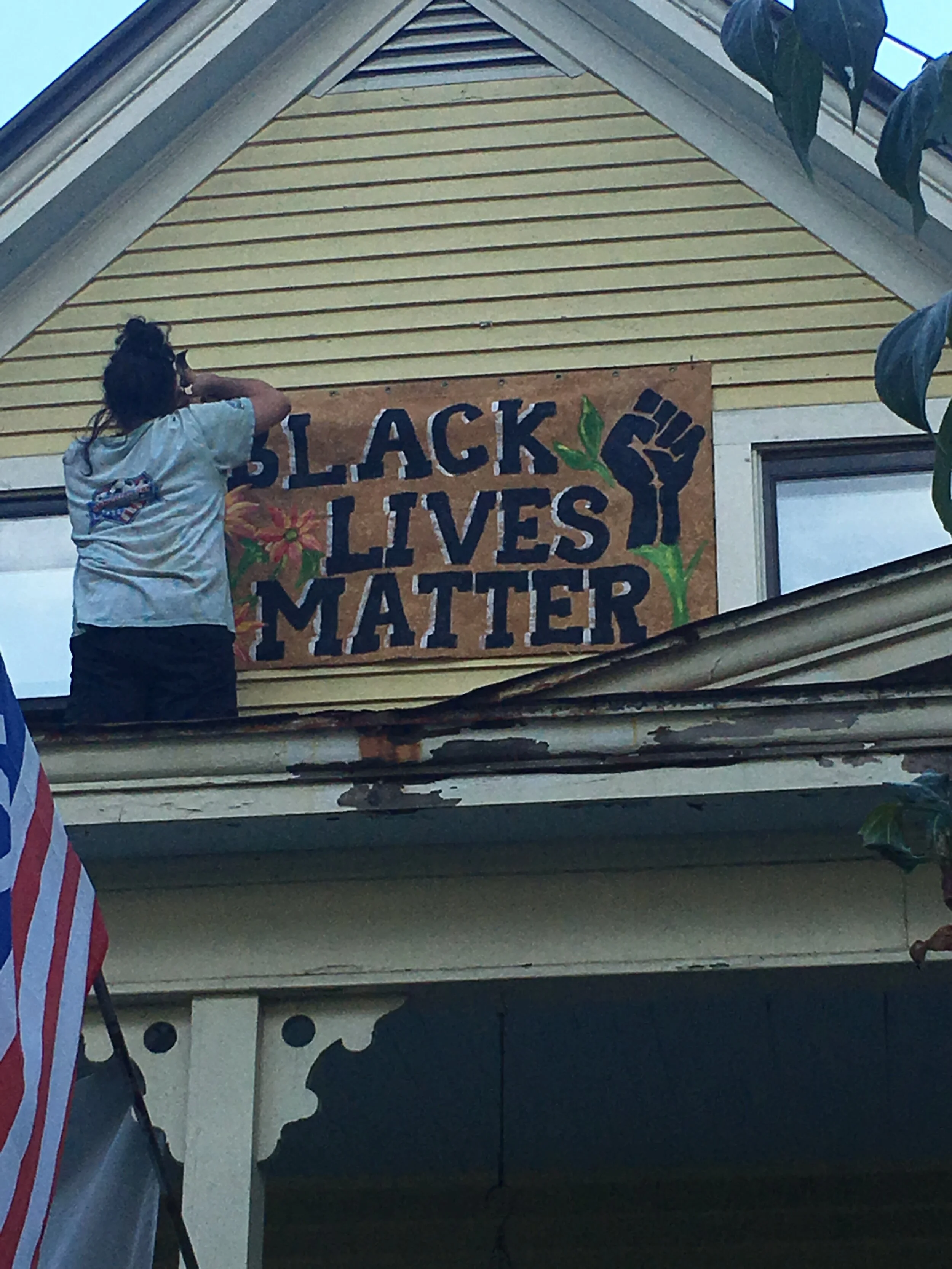 Person hanging large Black Lives Matter sign on second story of yellow house