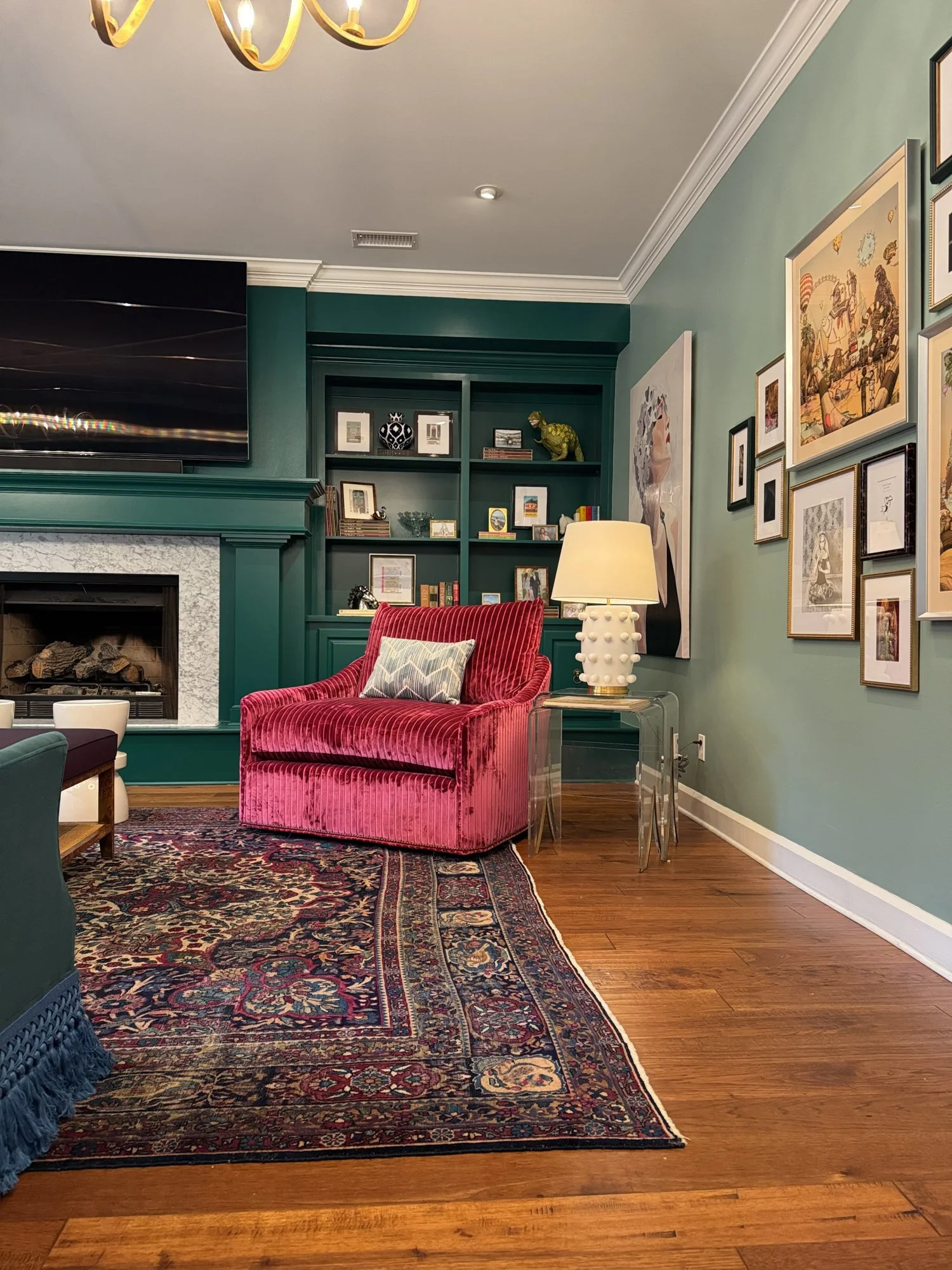 A corner of a green living room with a pink velvet armchair, a patterned rug, and built-in shelves