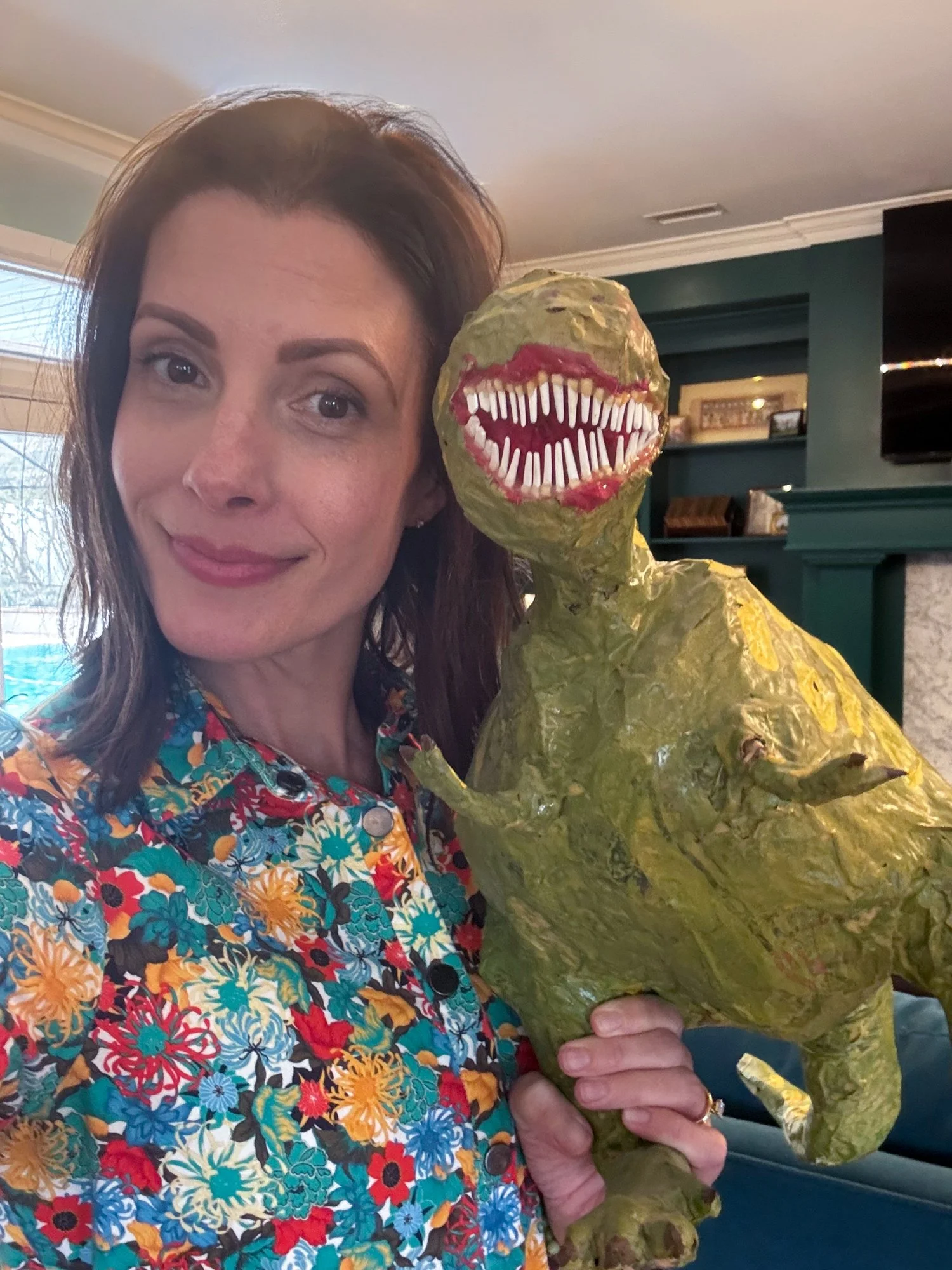 Lisa Gilmore holding up a handmade green paper-mâché dinosaur art piece