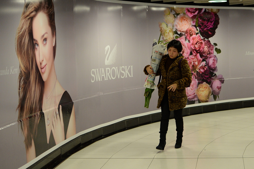 a woman carrying home some flowers for chinese new year passes in front of a huge swarovski billboard in the mtr.