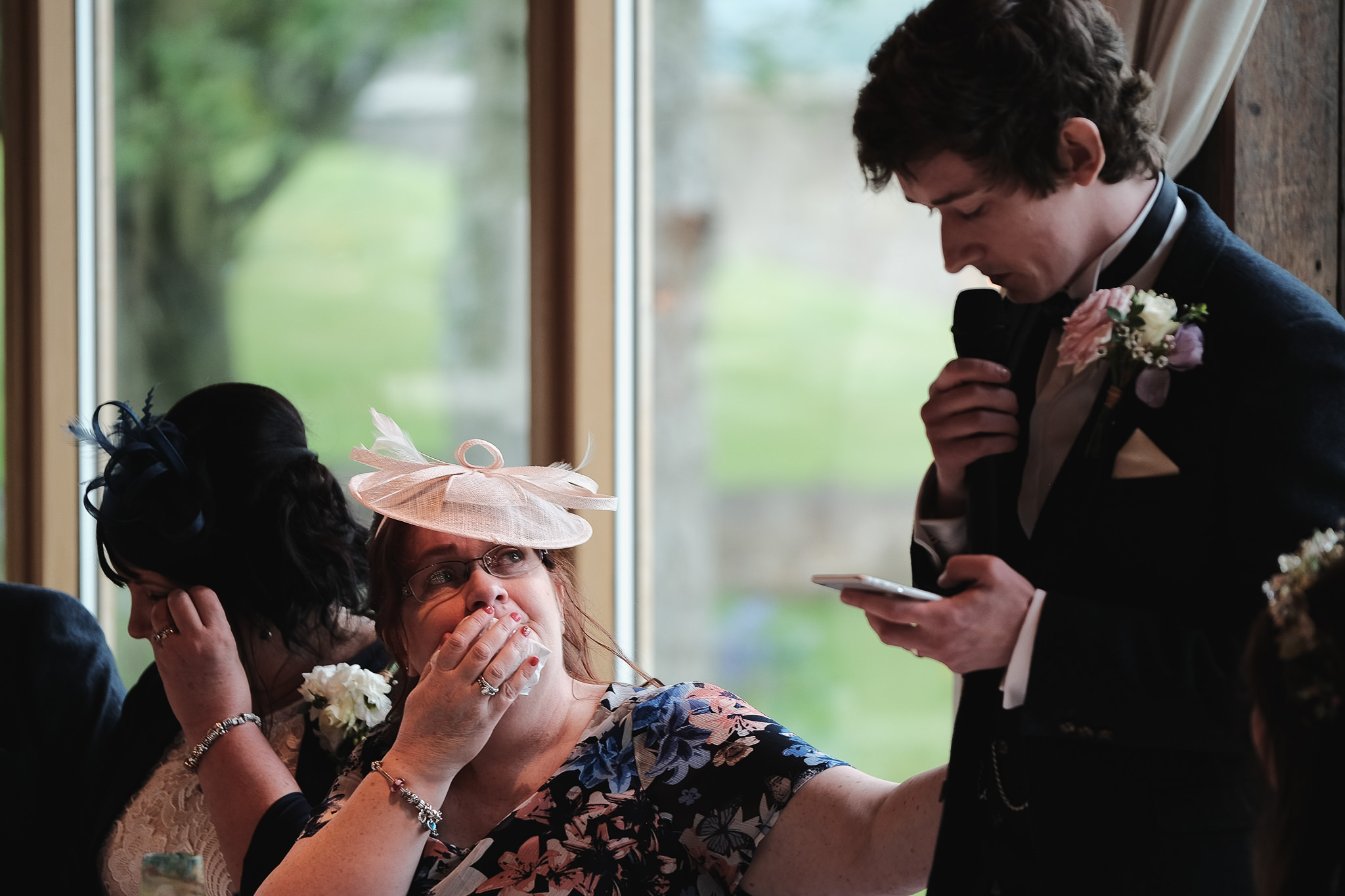 The crying mother of the groom reaches out to the groom as he cries while making his speech at Newton Hall, Northumberland by Barry Forshaw